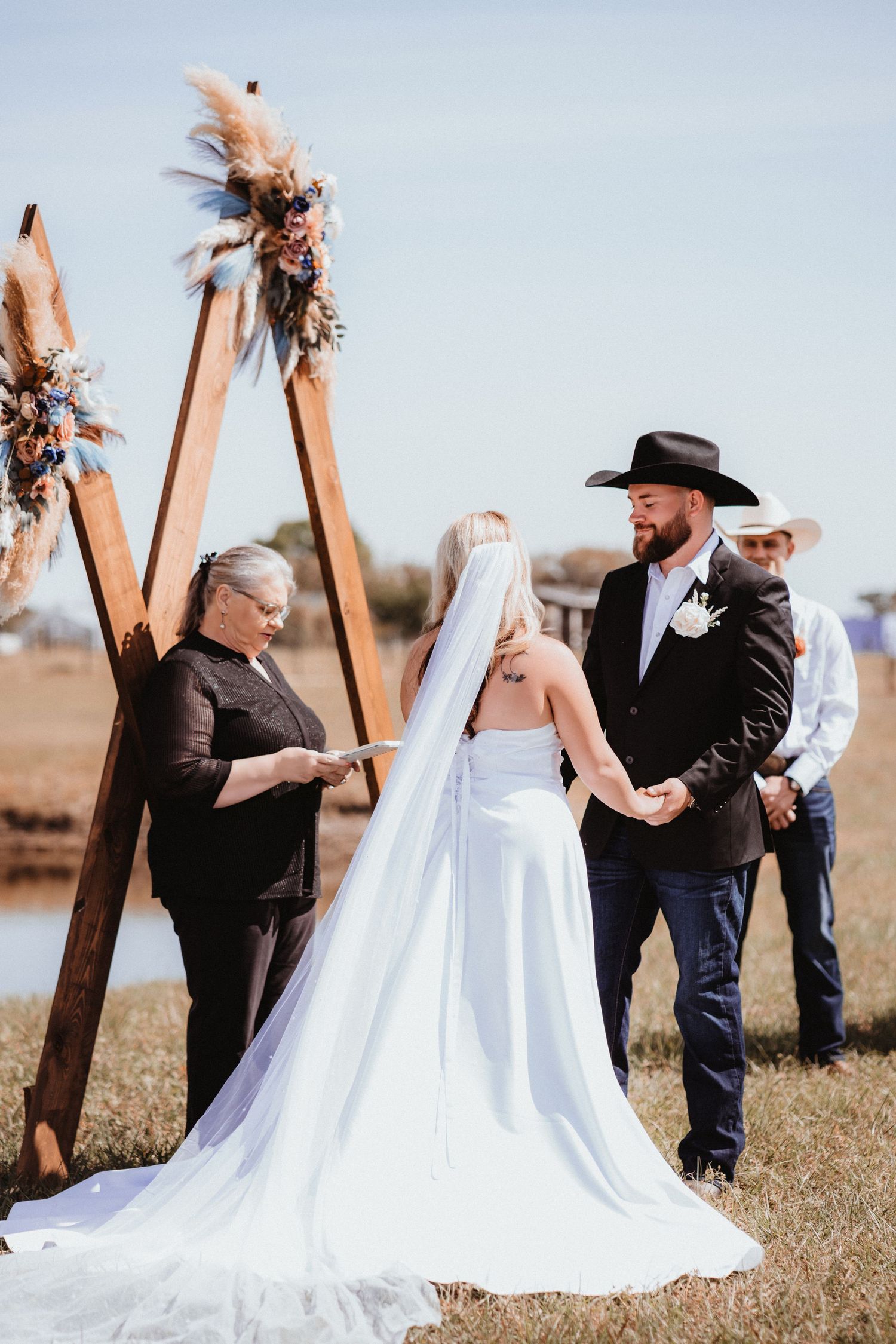 Bride and groom hold hands during outdoor wedding ceremony at a boho wooden arch as officiant reads vows.