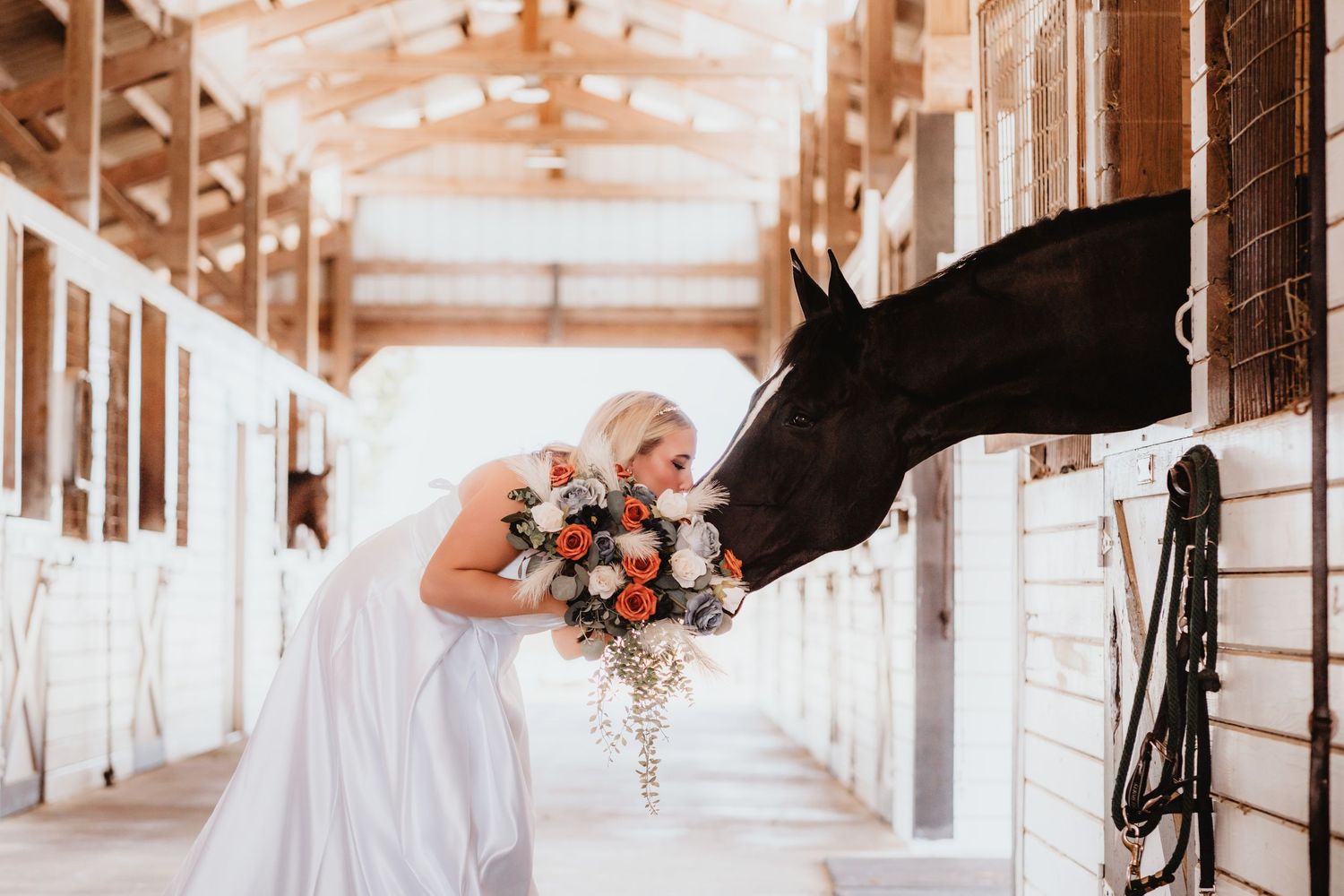 Bride in white gown leans toward a black horse in a rustic wooden stable aisle.