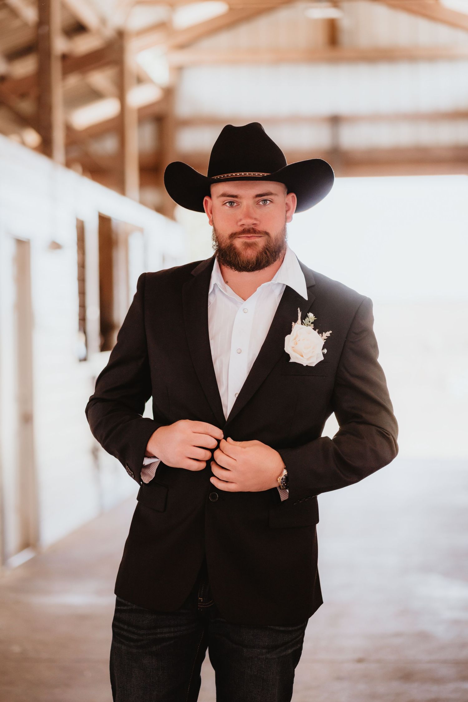 Confident groom in black cowboy hat adjusts blazer with white rose boutonniere in sunlit barn aisle.