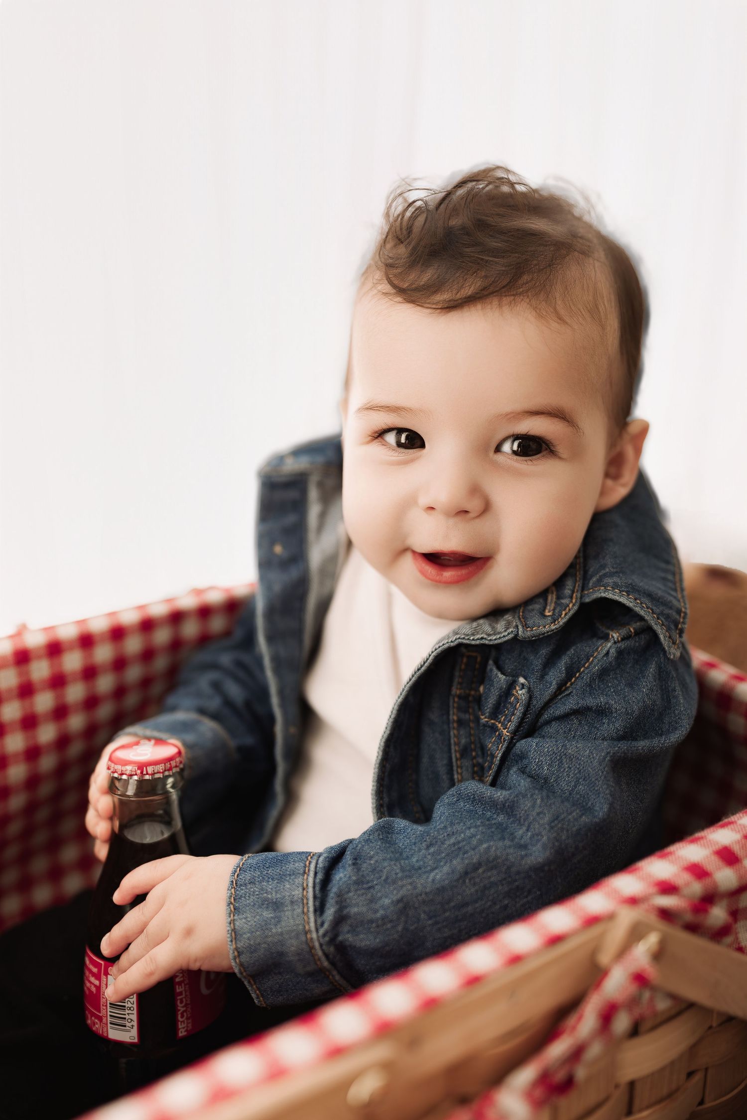 Smiling baby in denim jacket sitting in picnic basket holding a Coca-Cola bottle.
