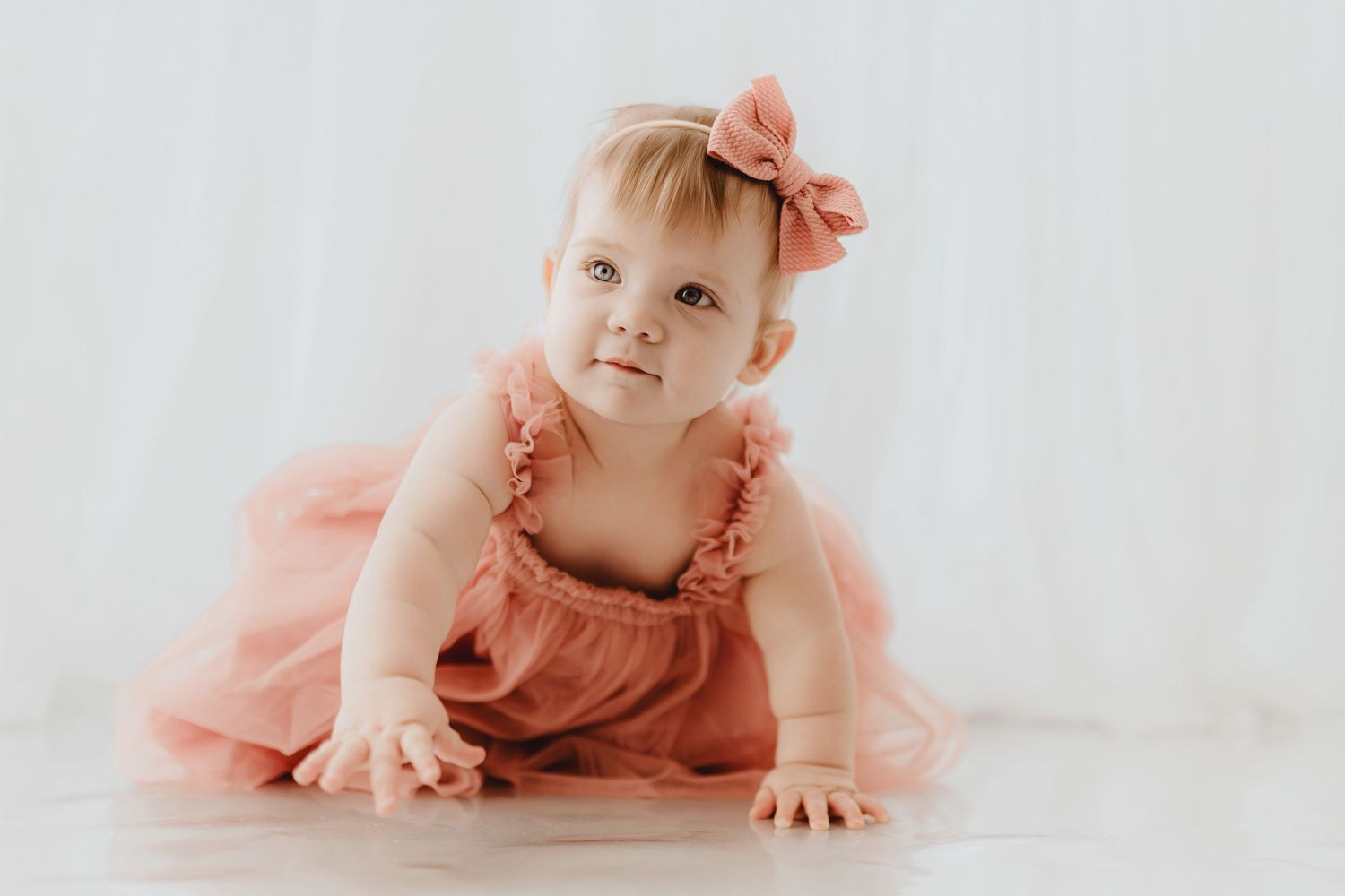 Baby girl in pink ruffle dress and bow headband crawling on white floor.