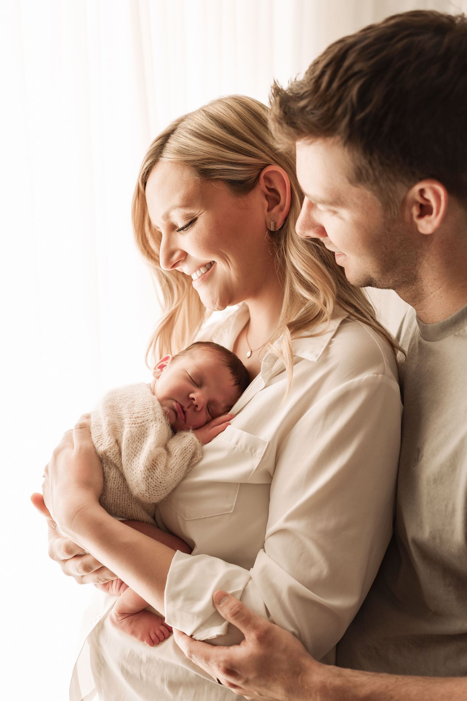 Happy parents smiling and holding their sleeping newborn baby by a bright window.
