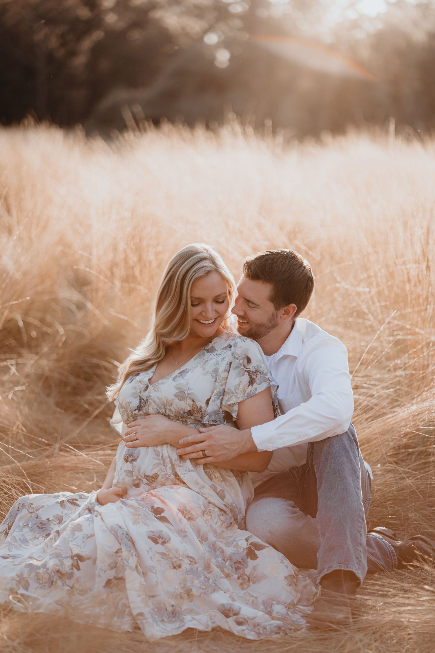 Couple sits in golden grass field, man tenderly holding pregnant woman's belly during maternity shoot.