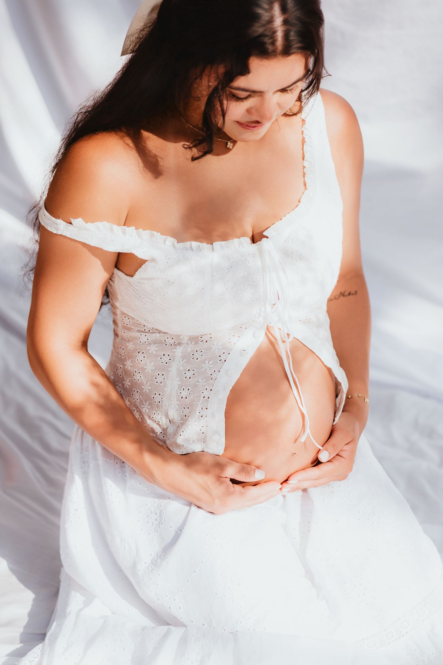 Close-up of pregnant woman in white lace dress gently cradling her baby bump.