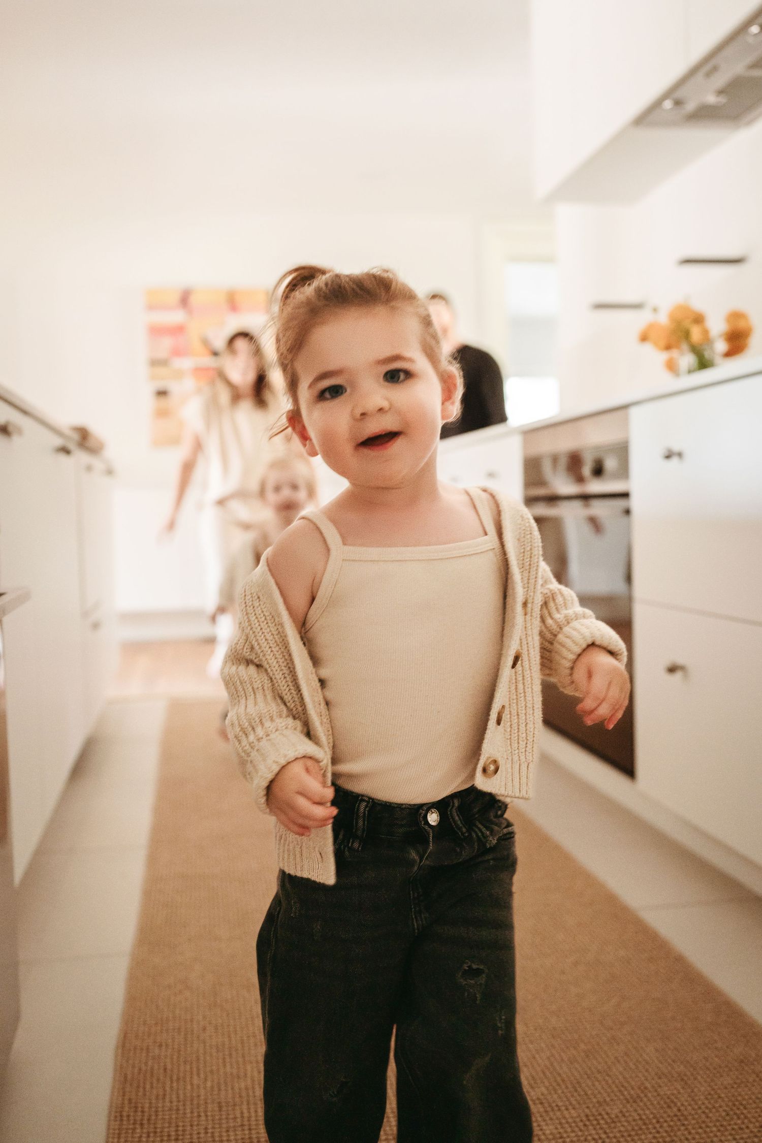 Adorable toddler girl in a cream cardigan smiling in a modern white kitchen.