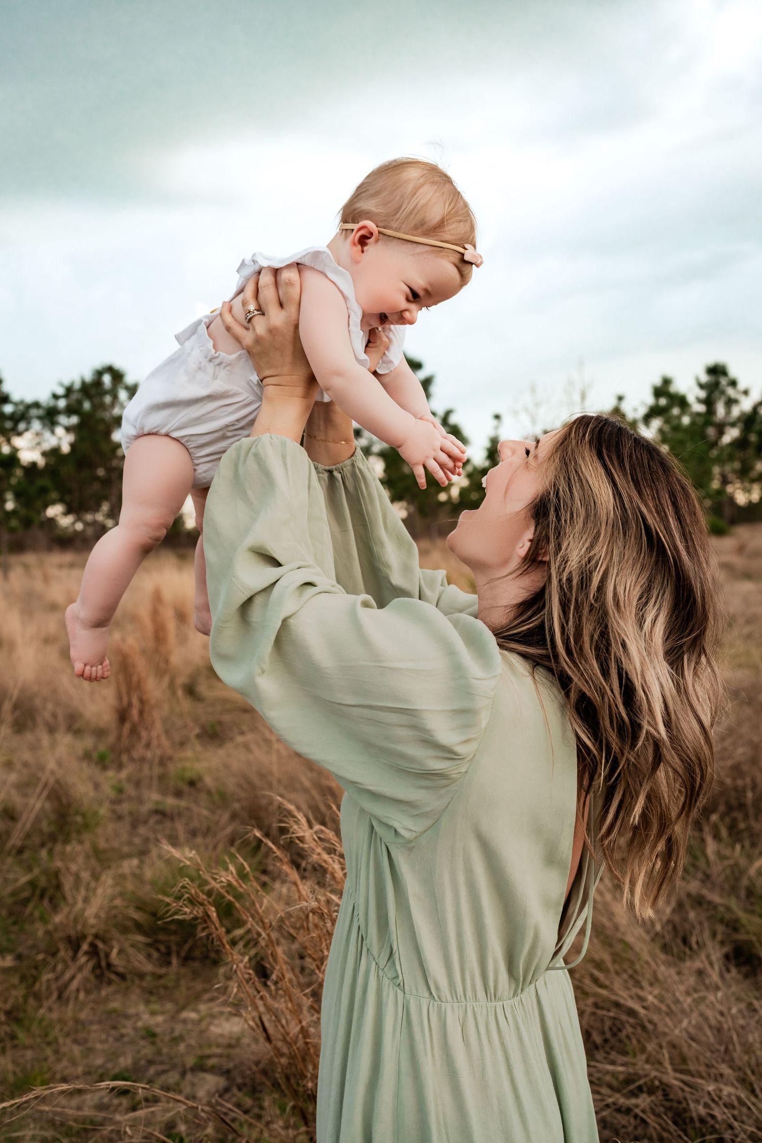 Mother in mint green dress lifting giggling baby girl overhead in a rustic outdoor field setting.