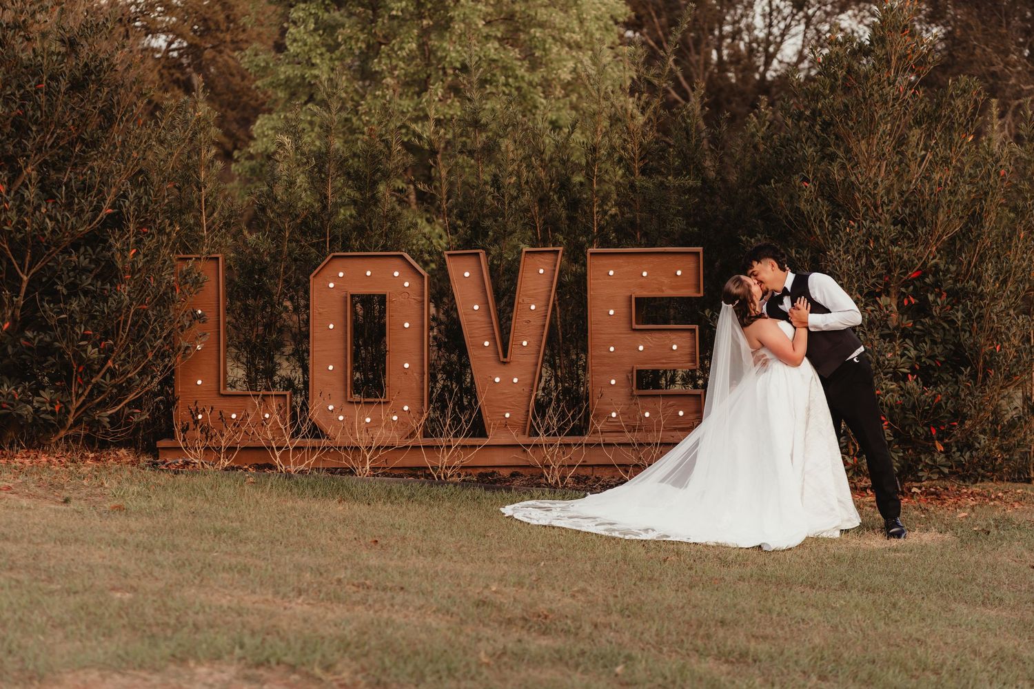 Bride and groom sharing a tender kiss in front of a glowing rustic LOVE marquee sign.