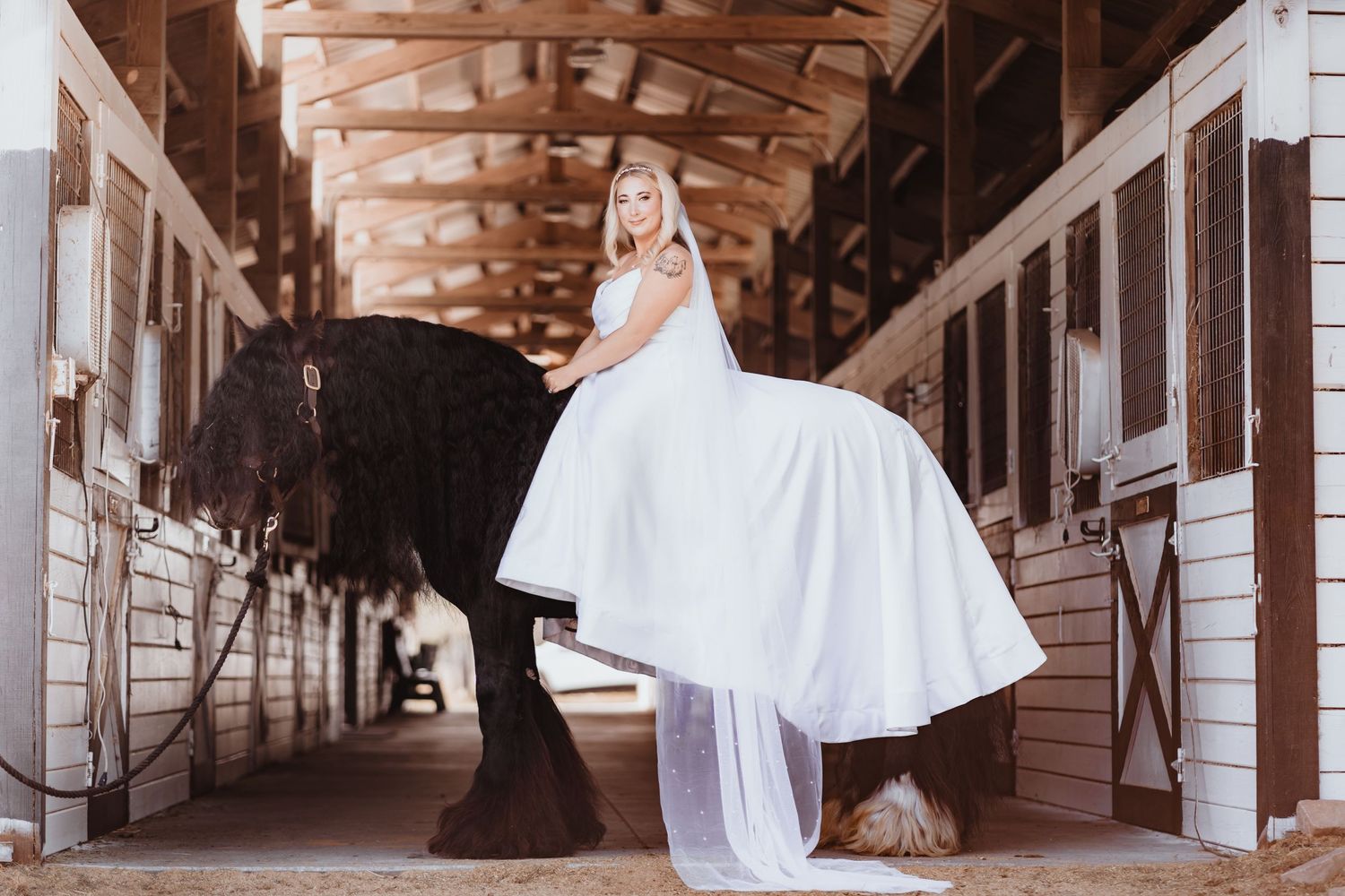 Bride in white gown sits elegantly on a black horse inside a barn stable.