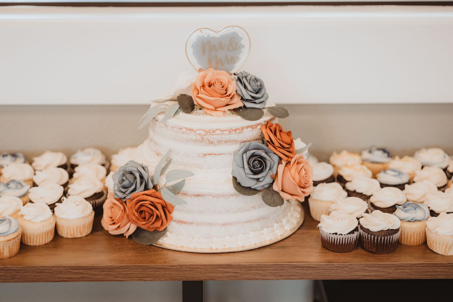 Two-tier semi-naked wedding cake decorated with orange and gray roses and heart topper, surrounded by cupcakes.