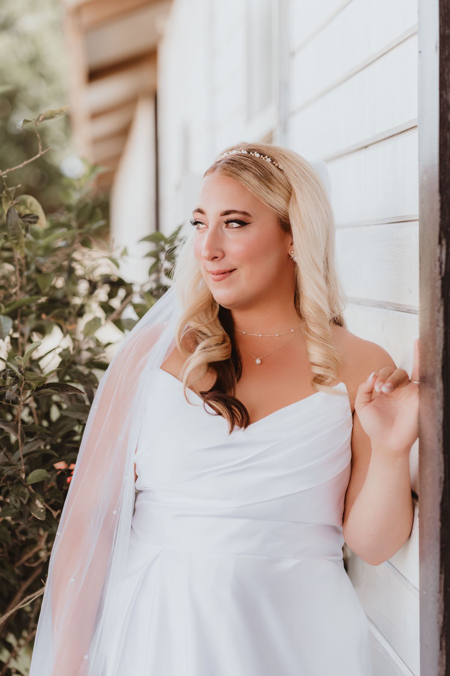 Beautiful blonde bride in strapless white gown with long veil and tiara leaning against white wooden wall outdoors.