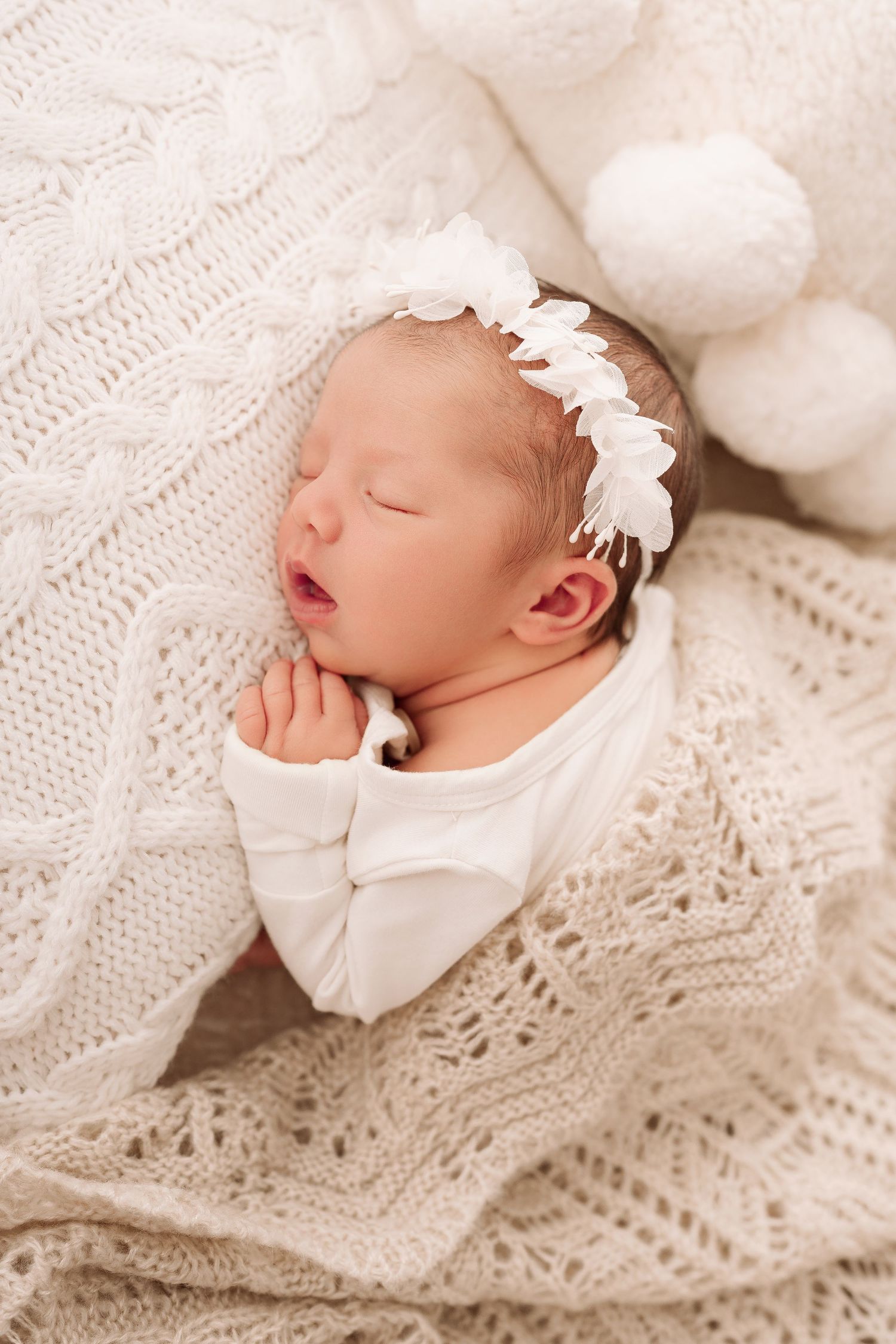 Sleeping newborn baby girl in white outfit and floral headband, wrapped in a cozy knit blanket.