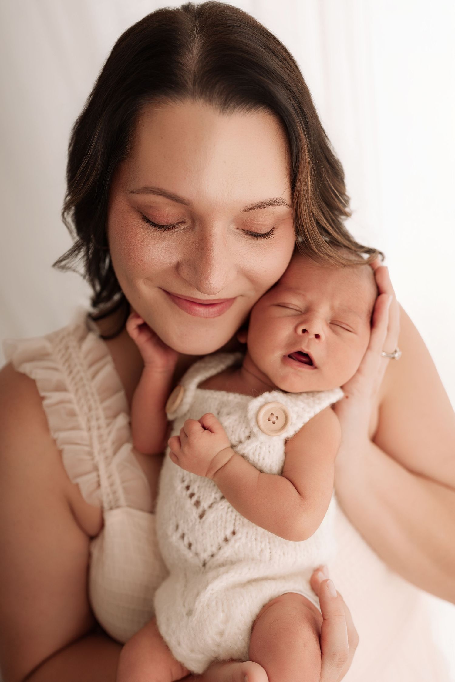 A smiling mother with closed eyes gently holds her sleeping newborn baby dressed in a white knit romper.