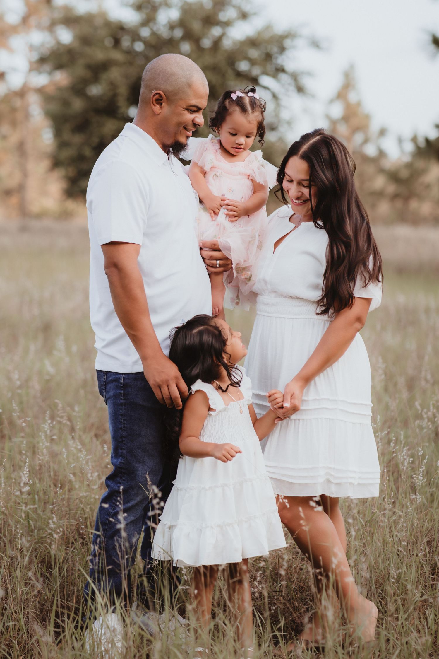 Happy family of four in white outfits posing together in a sunlit grassy field.