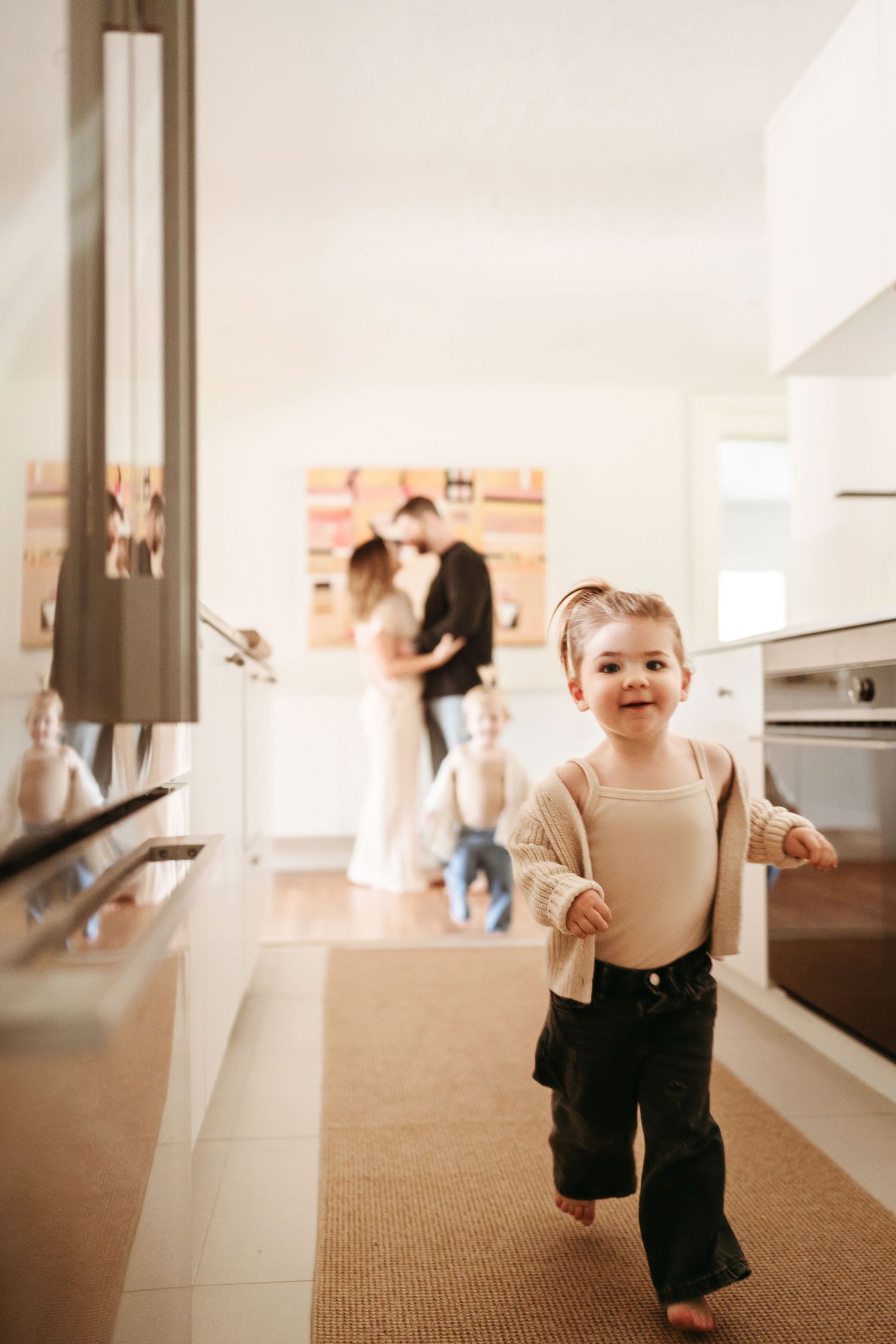A happy toddler runs through a modern white kitchen while parents embrace in the background.