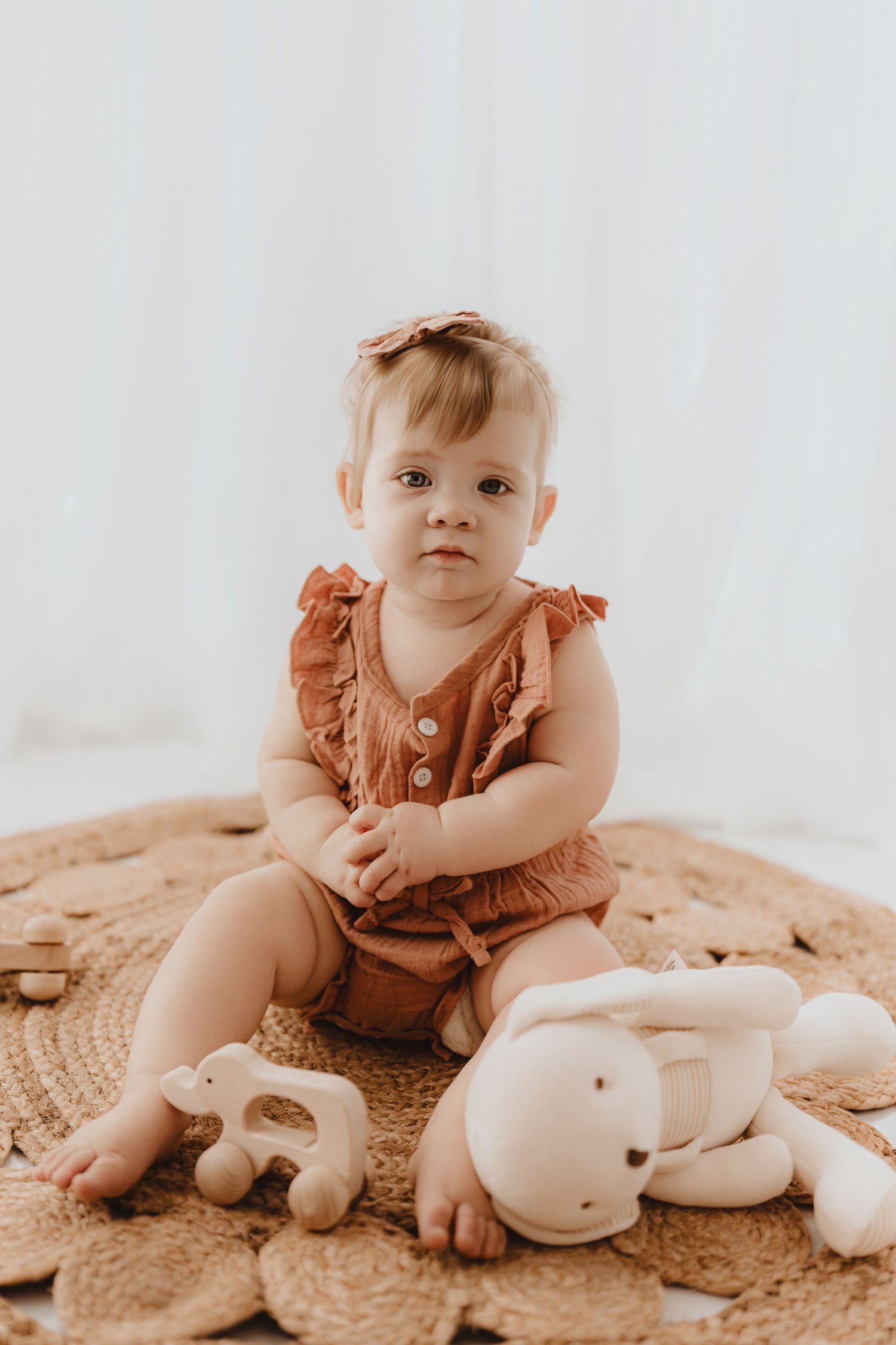 Cute baby girl in terracotta ruffle romper sitting on woven rug with plush bunny toy nearby.