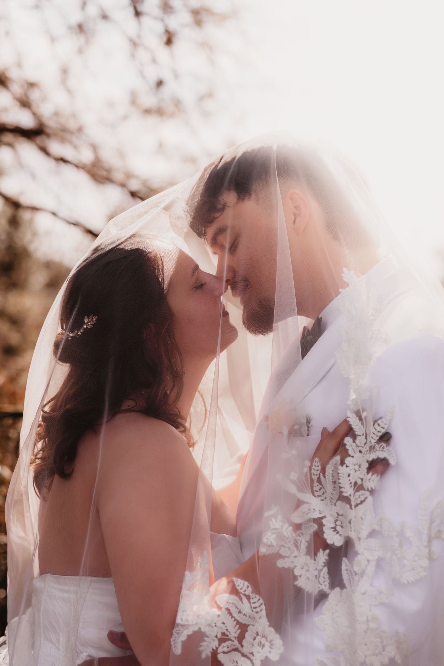 Romantic bride and groom almost kissing under a lace wedding veil in warm backlit sunlight.