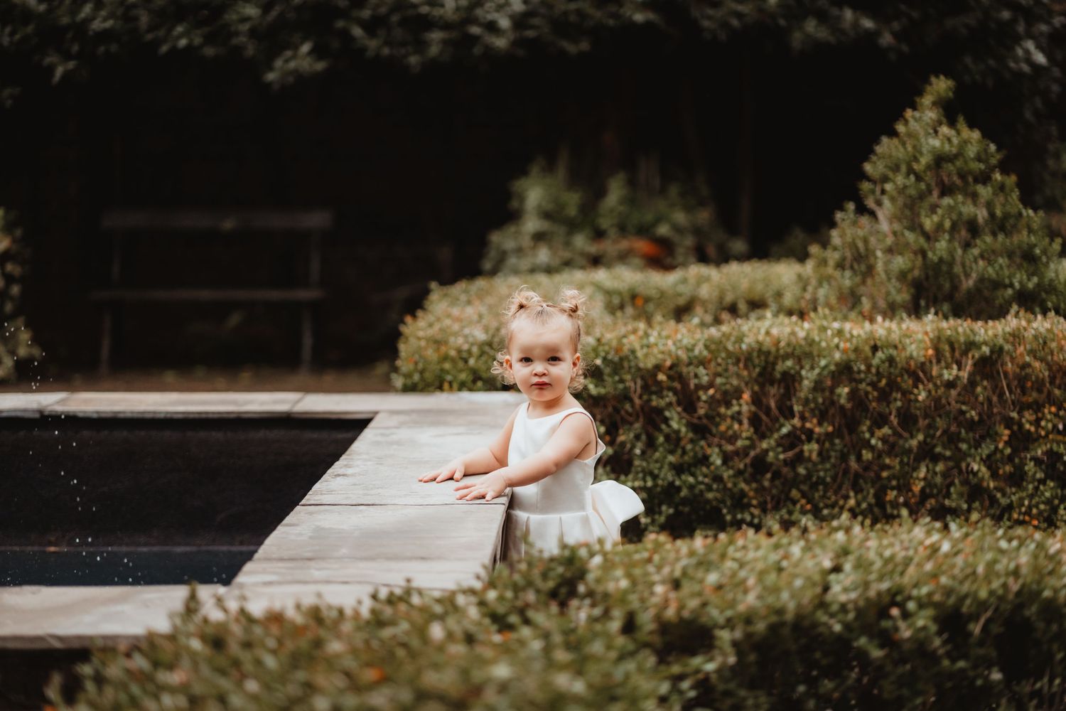 Adorable toddler in white dress stands by a garden pool surrounded by manicured hedges.