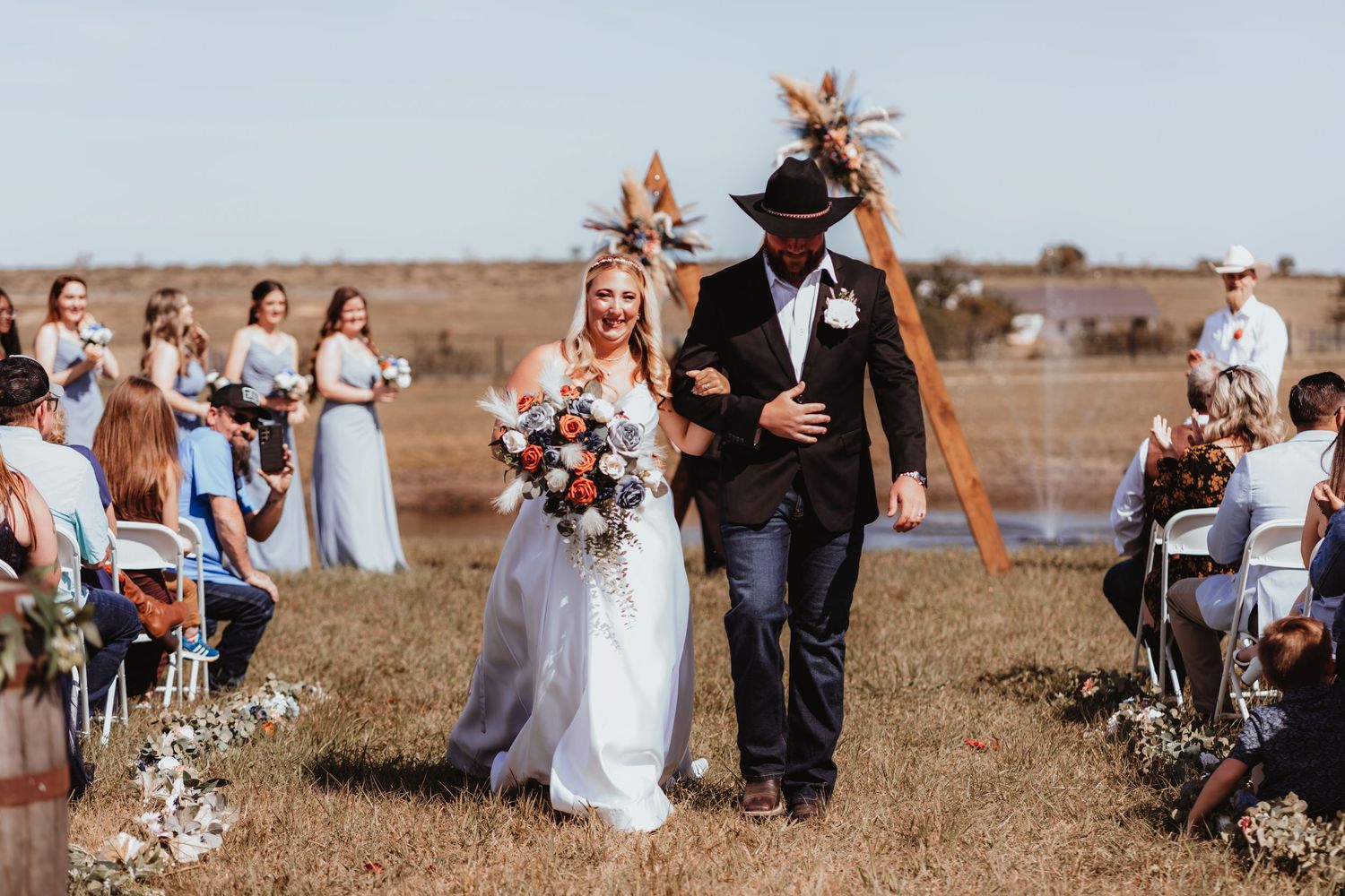 Bride and groom in cowboy hat walk down grassy aisle past guests at rustic outdoor wedding ceremony.