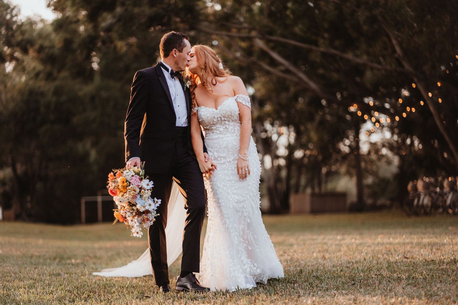 Newlyweds share a tender kiss outdoors at golden hour, bride in lace mermaid dress, groom in classic black tuxedo.