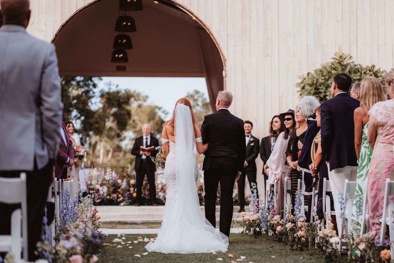 Bride and escort walk down outdoor wedding aisle toward arch with guests seated on both sides.