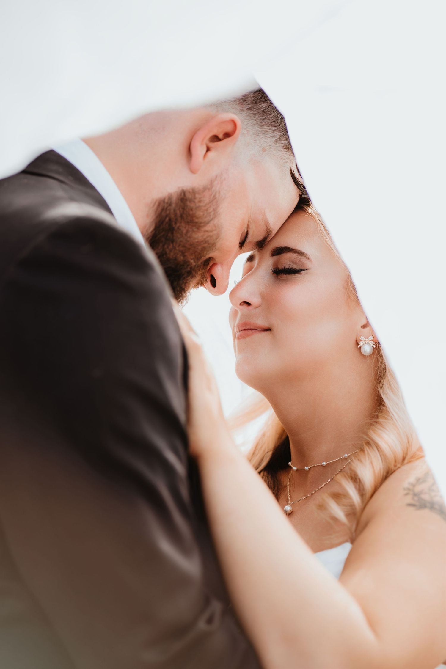 Groom and bride touch foreheads in an intimate moment under a veil, bride wearing pearl earrings and necklace.