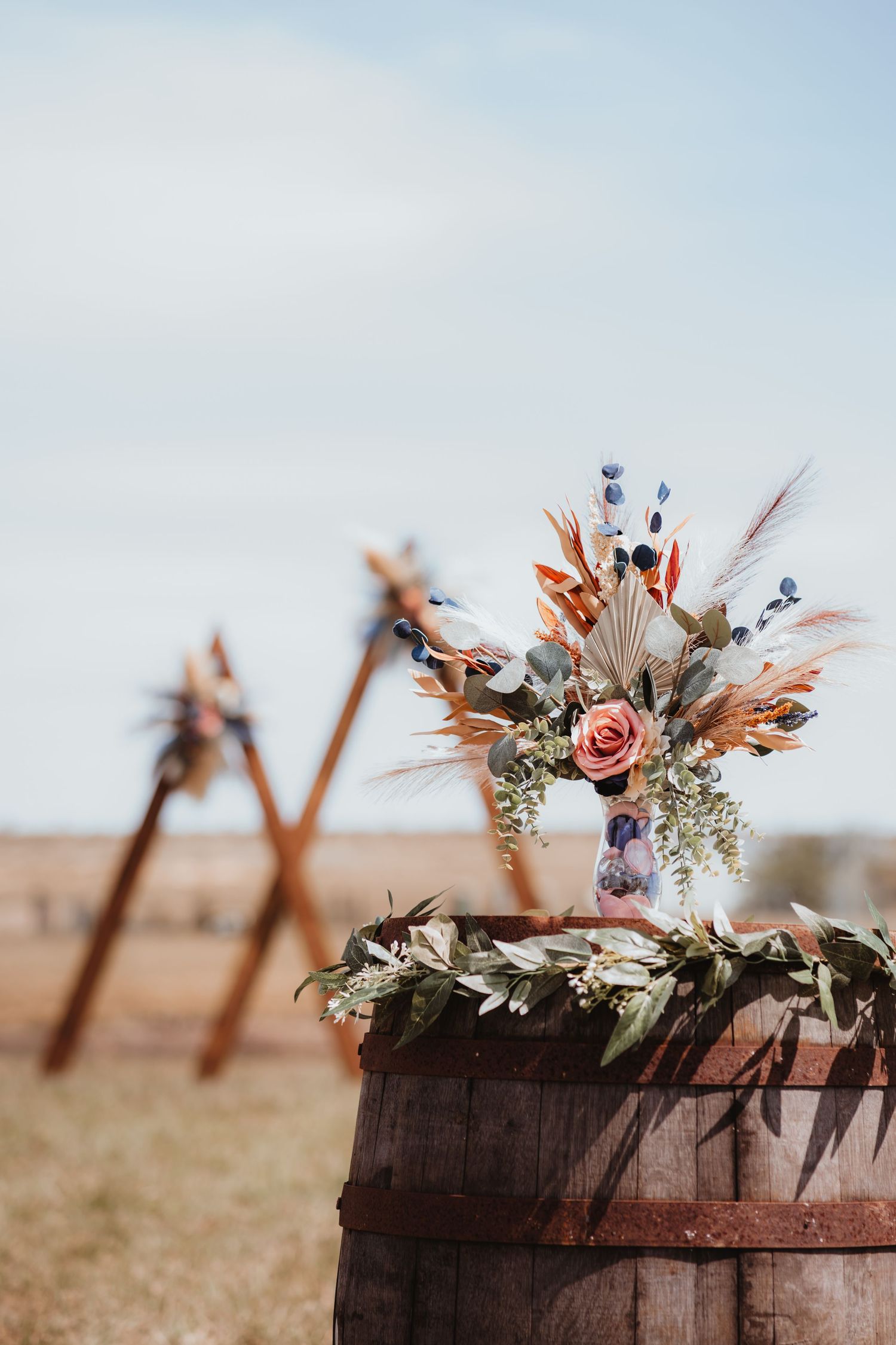 A boho floral arrangement on a rustic barrel at an outdoor wedding venue.