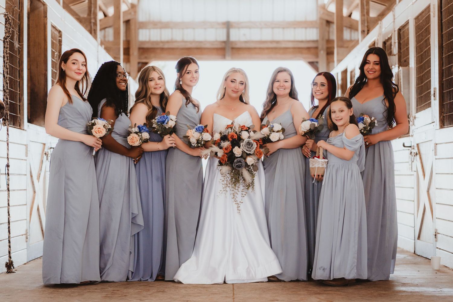 Bride in white gown poses with bridesmaids in dusty blue dresses inside a rustic barn venue.