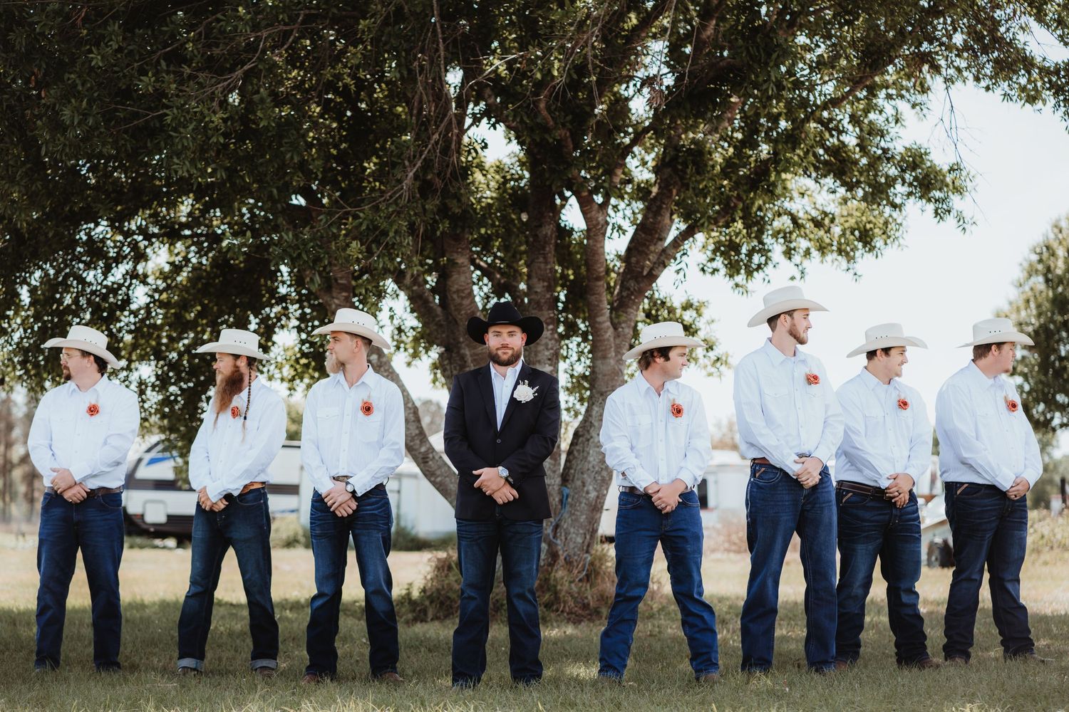 Groom in black cowboy hat stands with groomsmen in white shirts under large tree outdoors.