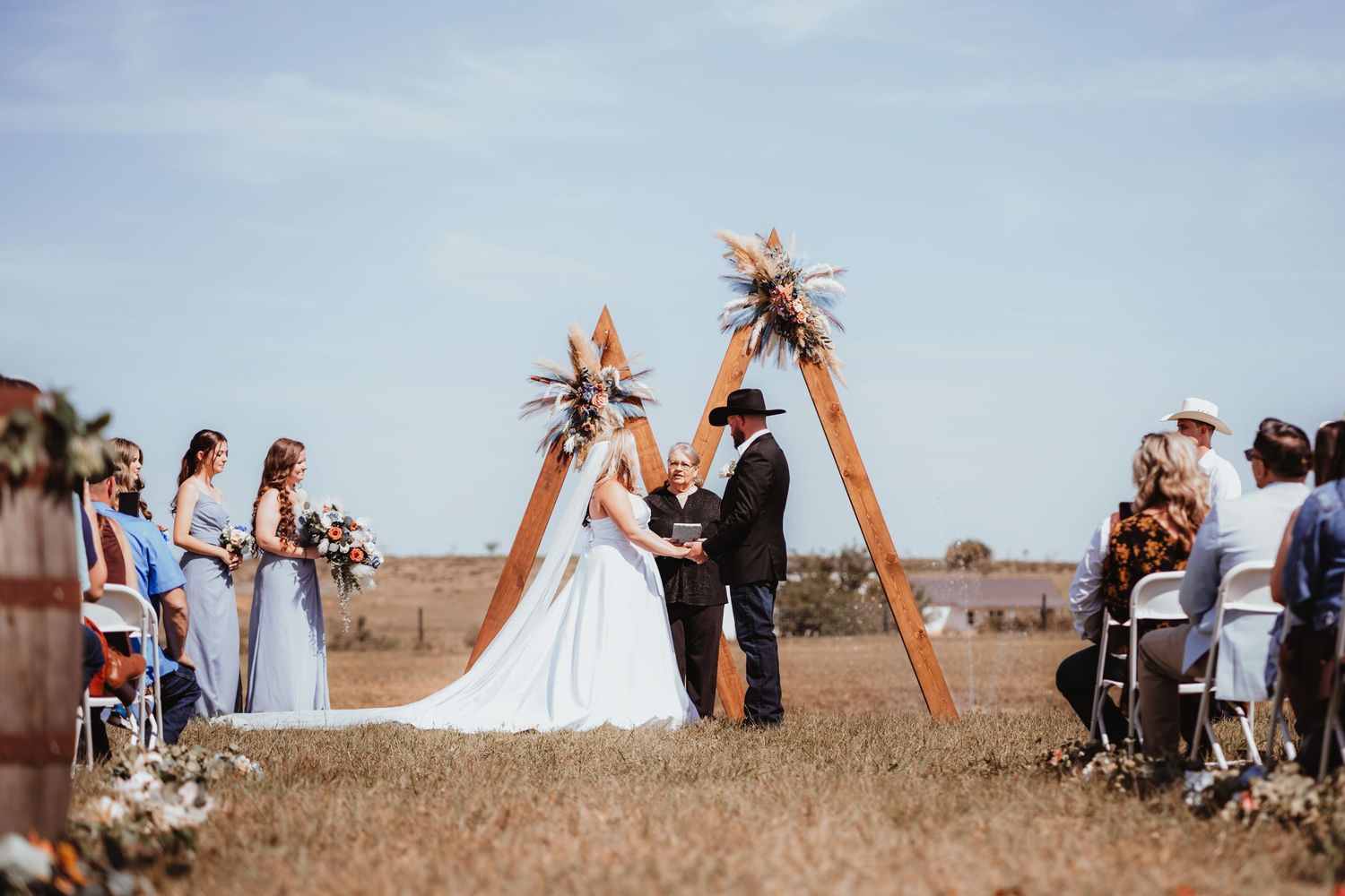 Bride and groom exchange vows under a wooden triangular arch at a rustic outdoor field wedding ceremony.