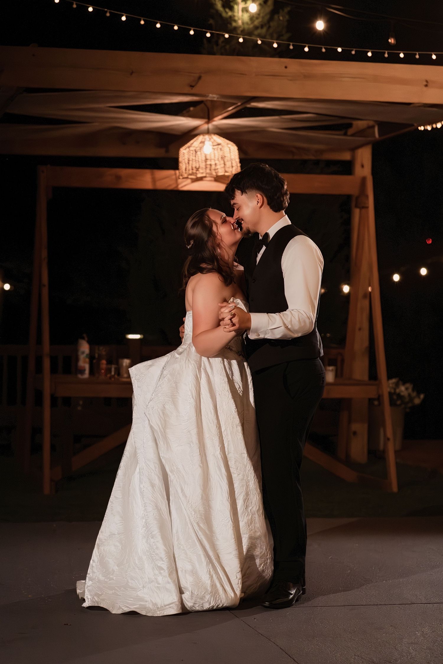 Newlyweds share an intimate moment during first dance under chandelier and string lights at night wedding.