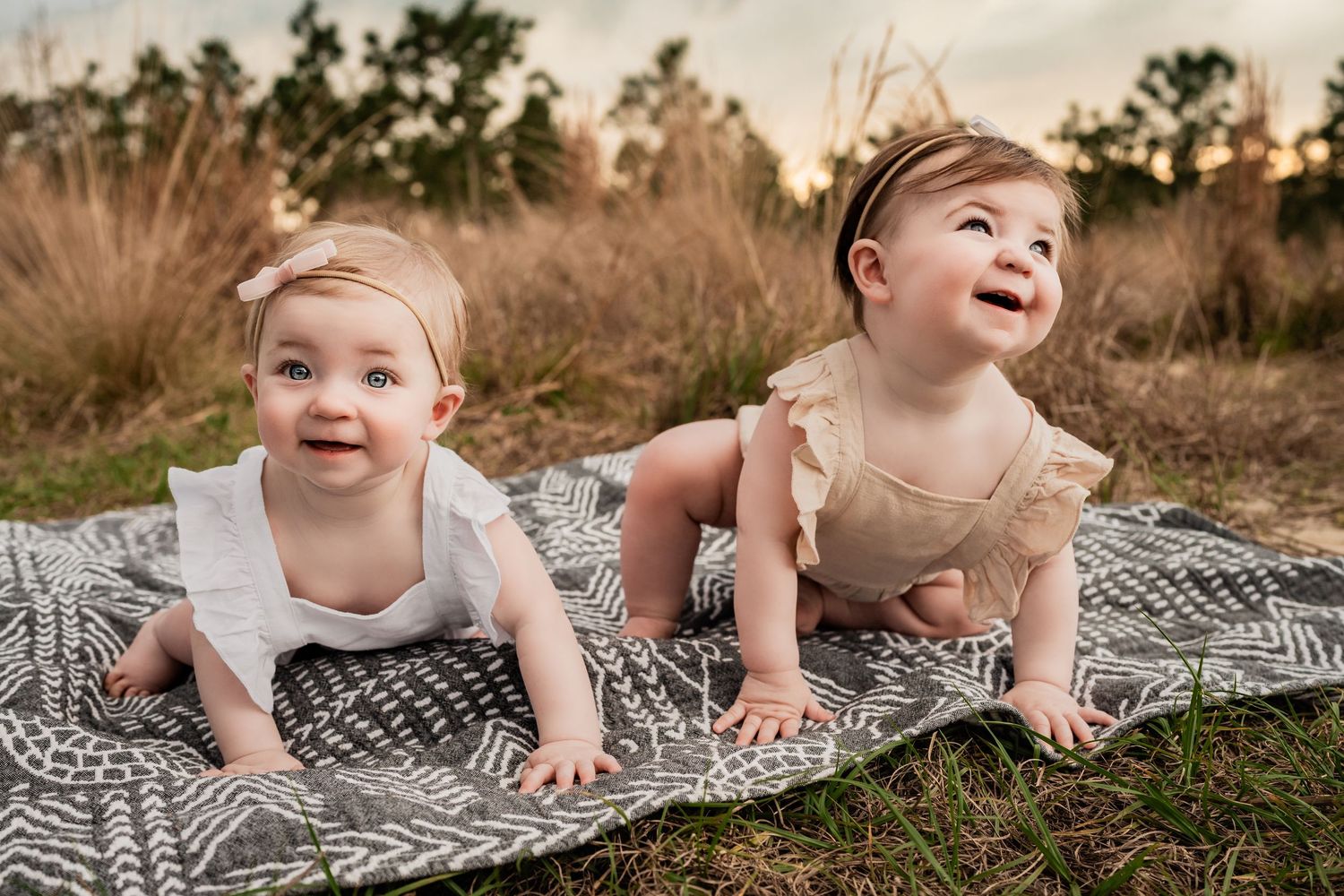 Two smiling baby girls crawling on a patterned blanket outdoors in a golden grass field at sunset.
