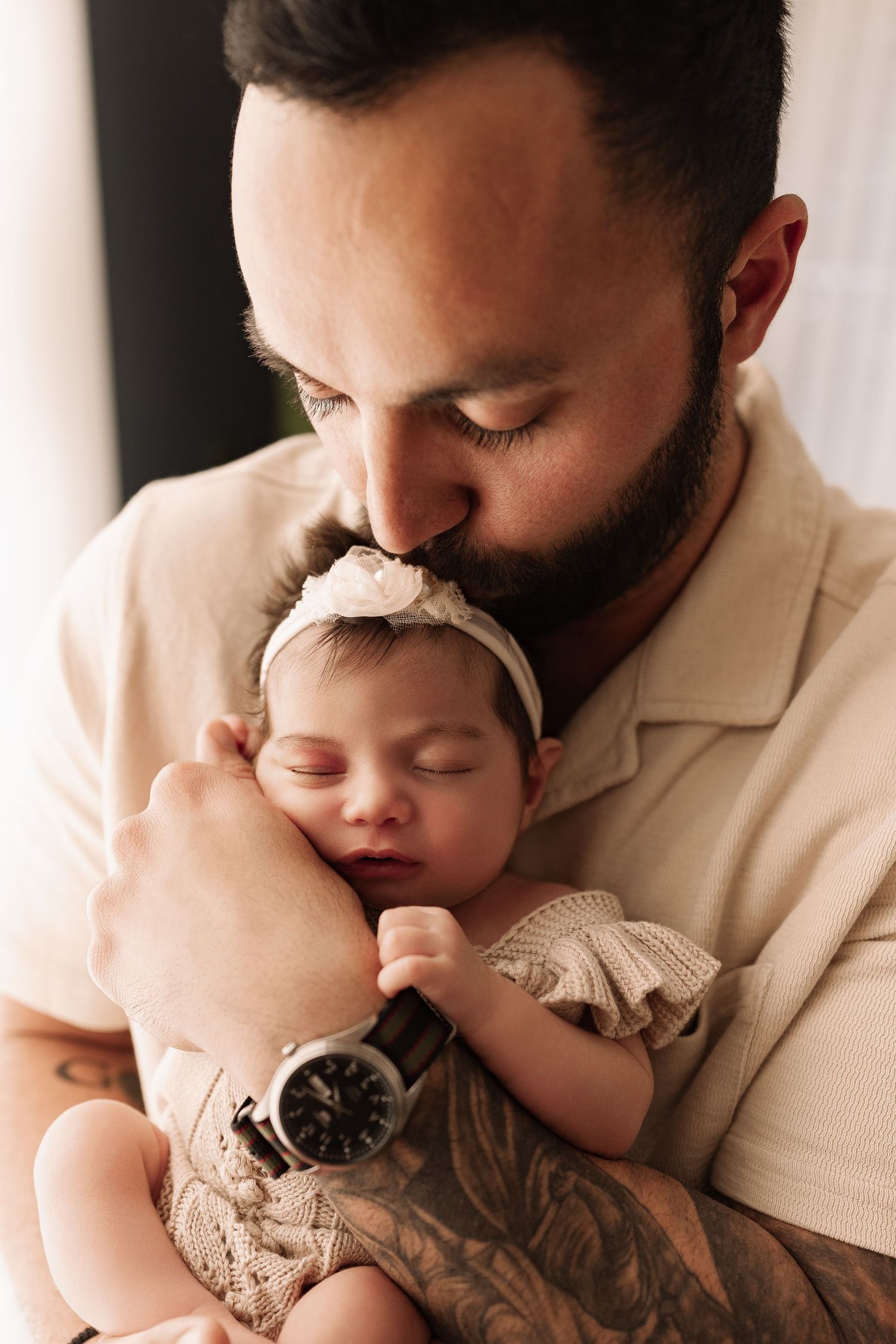 Bearded tattooed father gently kissing sleeping newborn baby girl wearing a white floral headband.