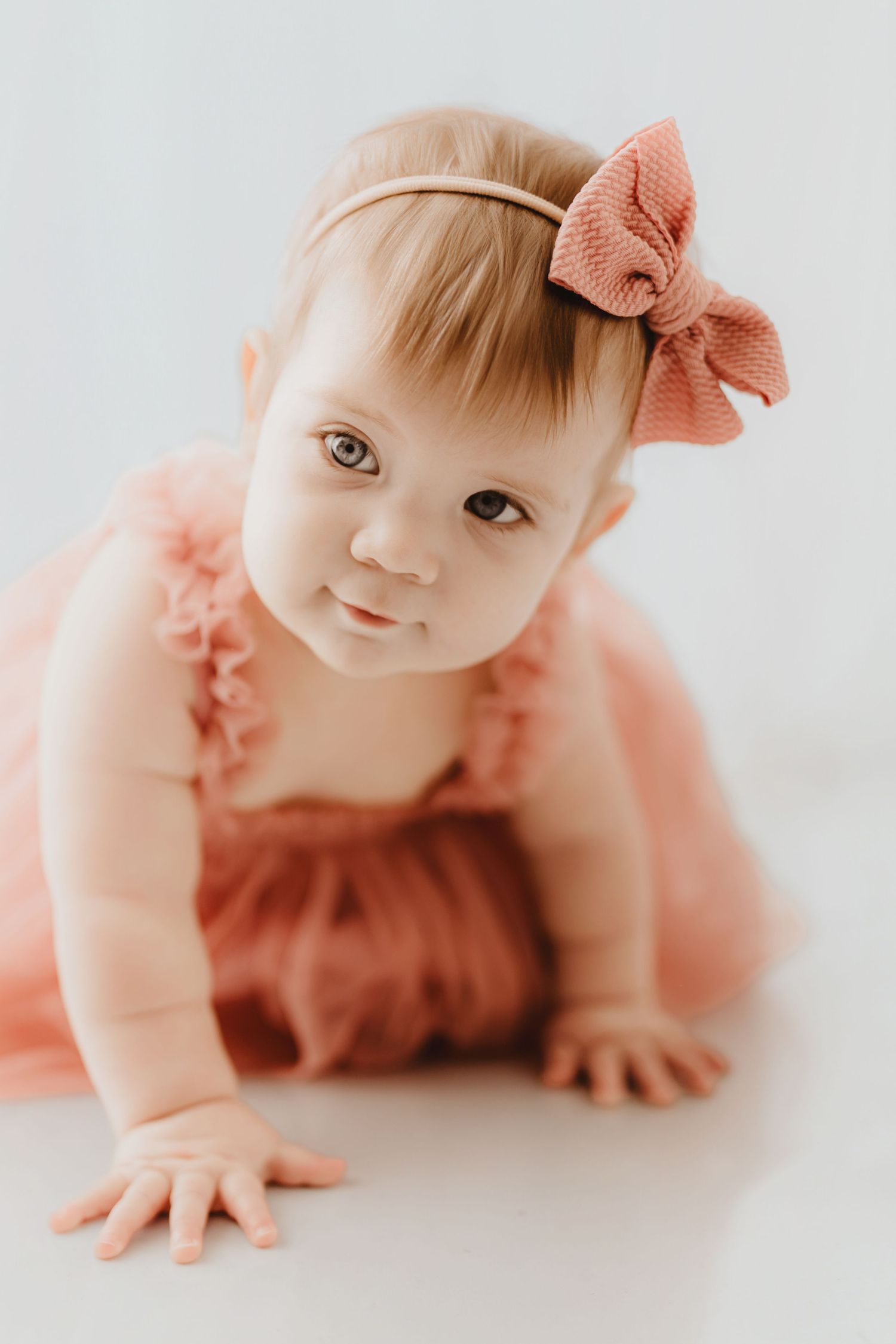 Adorable baby girl in coral tulle romper with matching bow headband looking at camera while crawling.