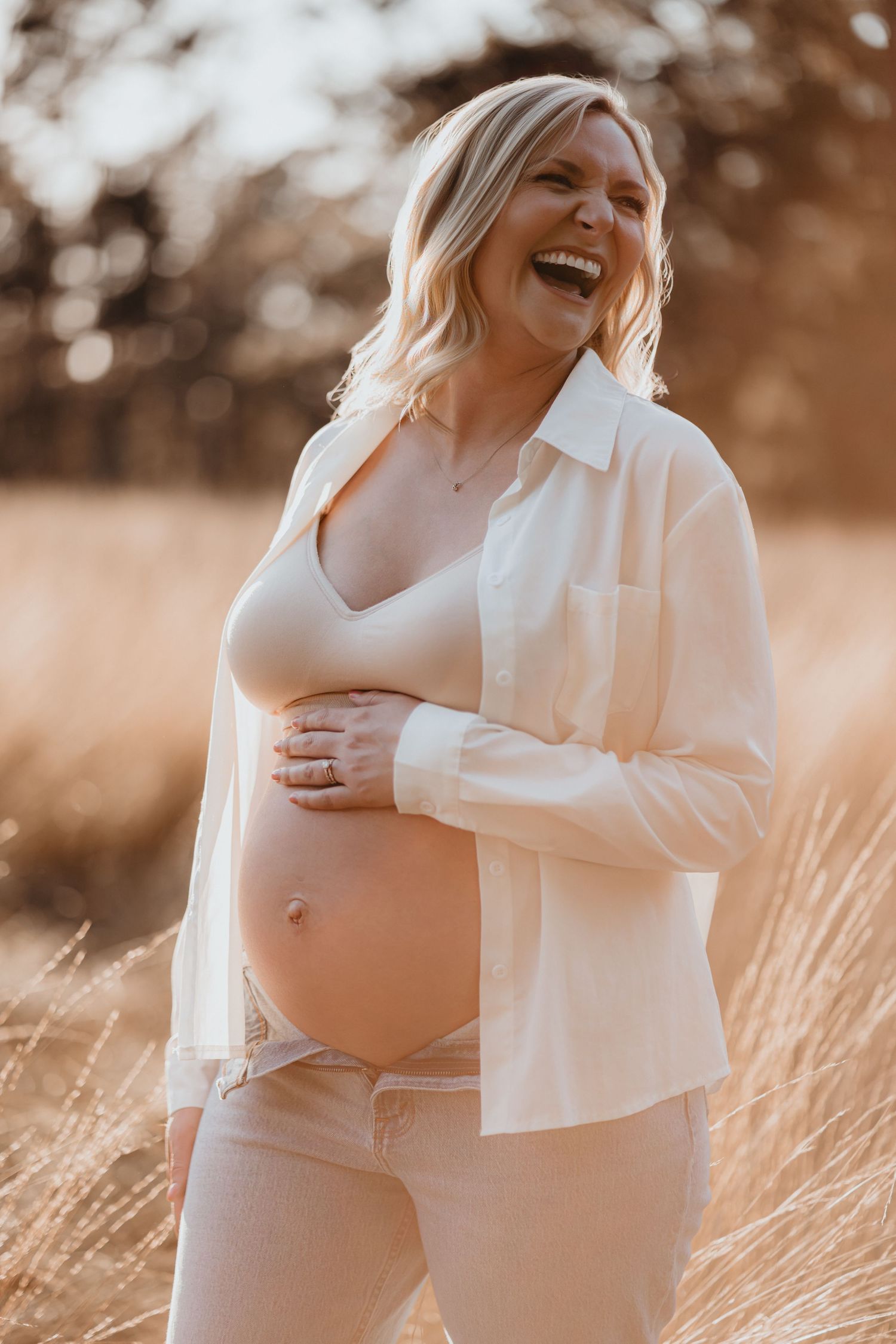 Joyful pregnant woman in white shirt laughs while holding bare belly in warm sunlit grass field.