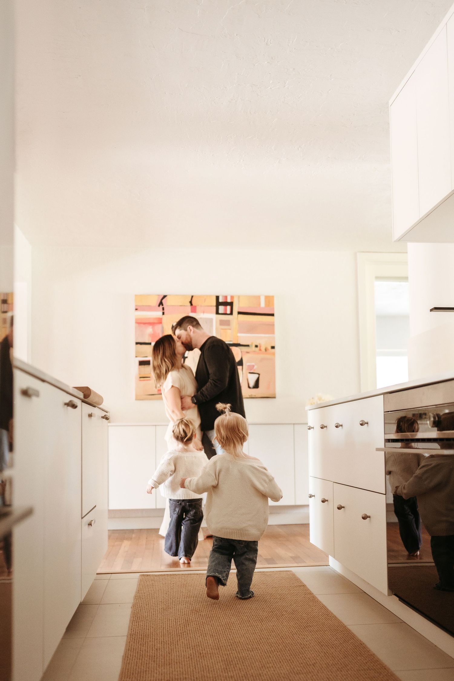 A couple shares a tender kiss in a white kitchen while two young children run toward them.