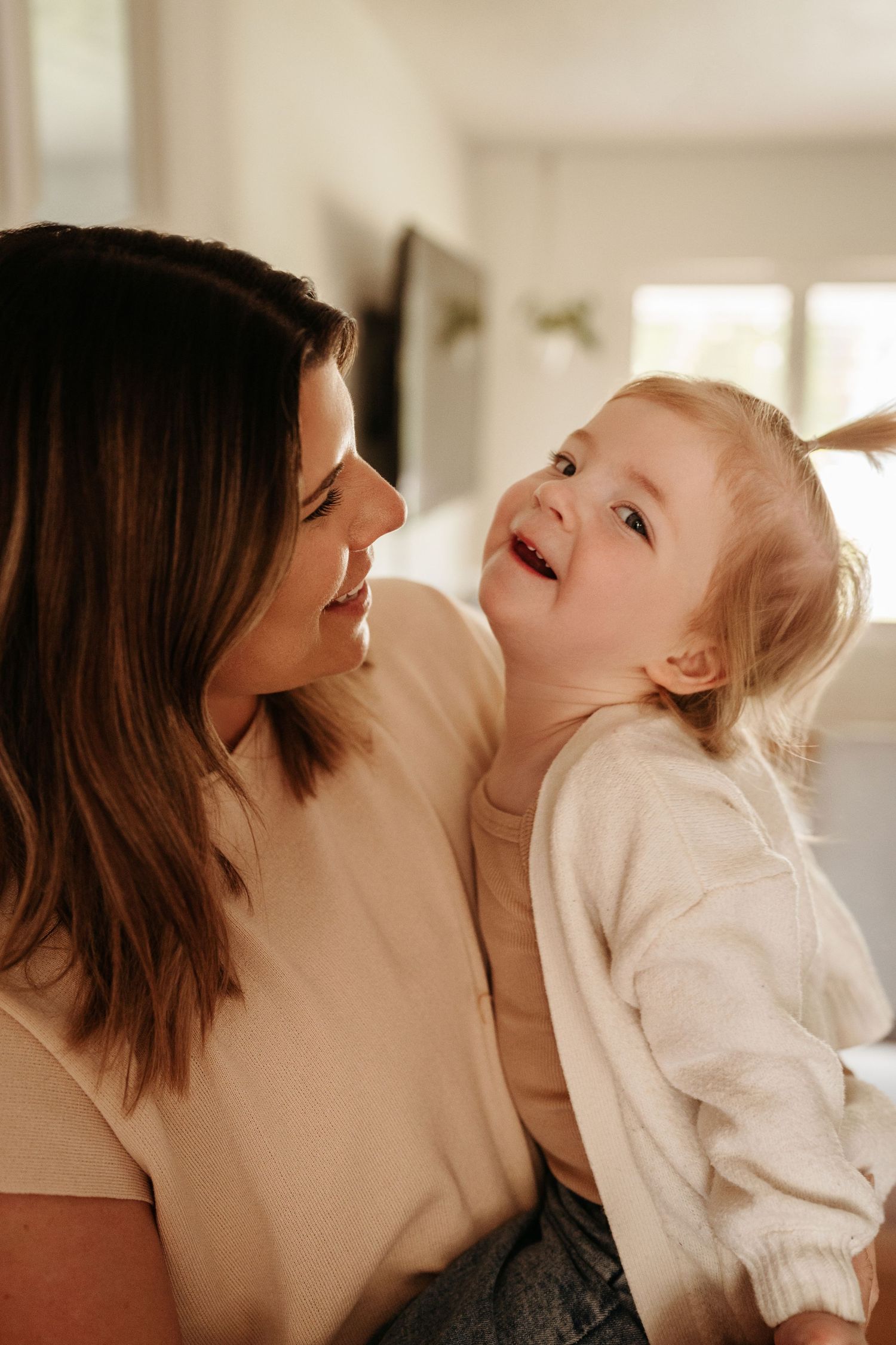 A brunette mother smiles lovingly at her laughing blonde toddler daughter in a bright home.