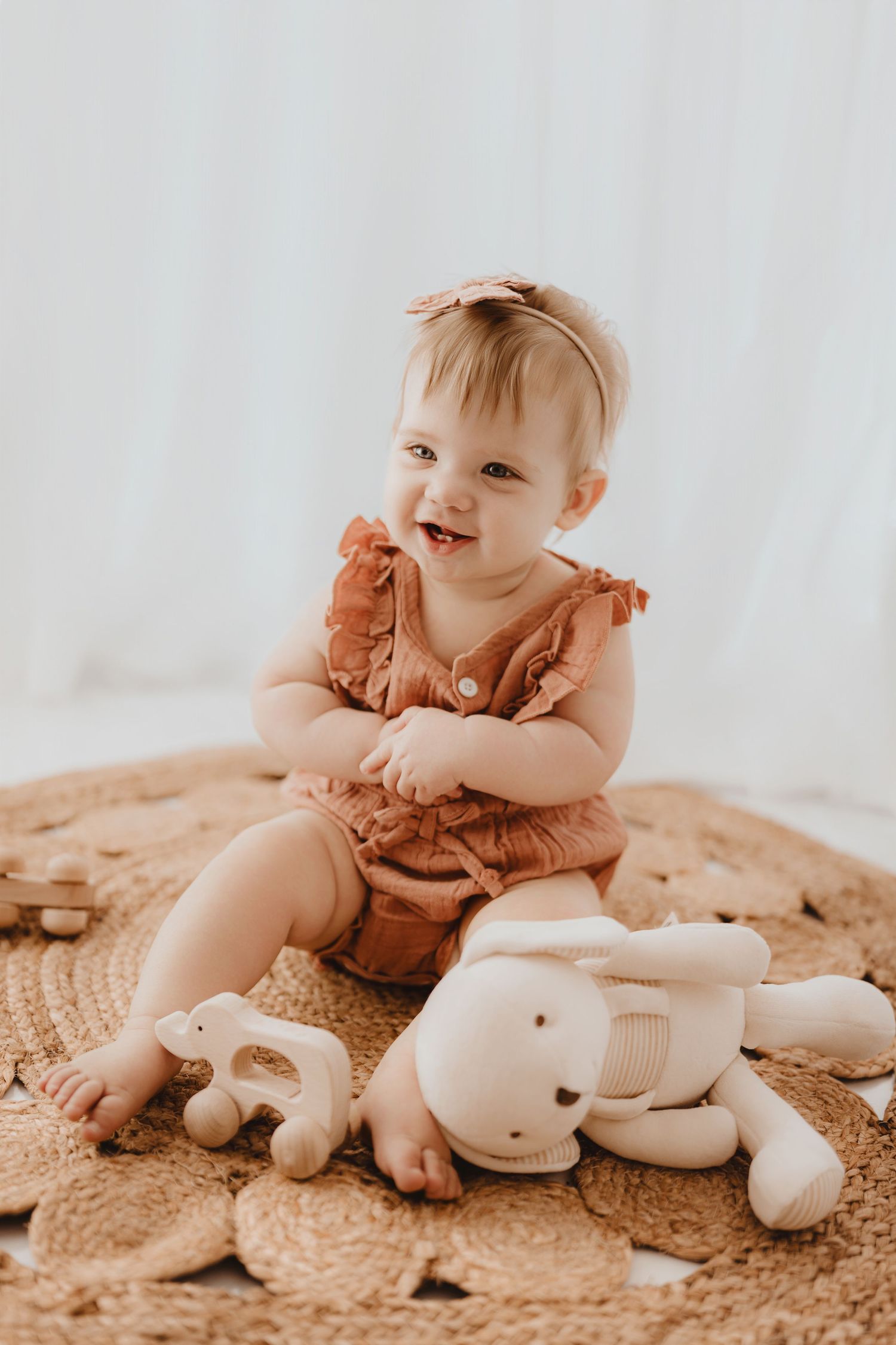 Happy baby girl in rust romper laughing on woven rug with stuffed bunny and wooden toys.