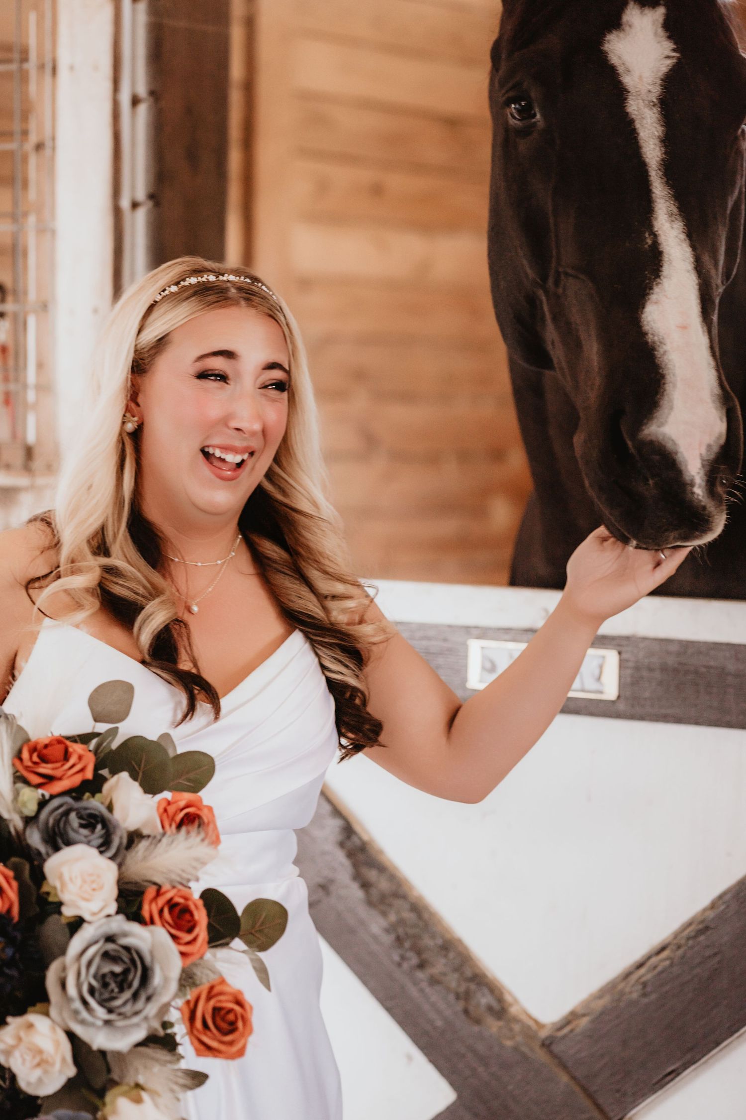 Joyful bride in white dress laughs while petting a black and white horse in stable.