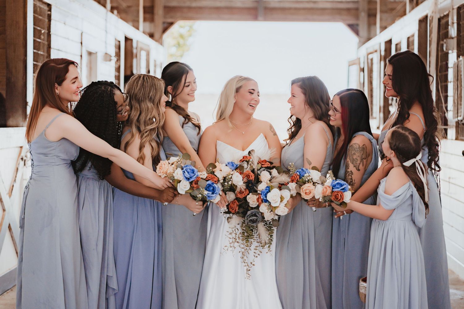 Bride in white gown holds colorful bouquet surrounded by bridesmaids in dusty blue dresses in a barn setting.