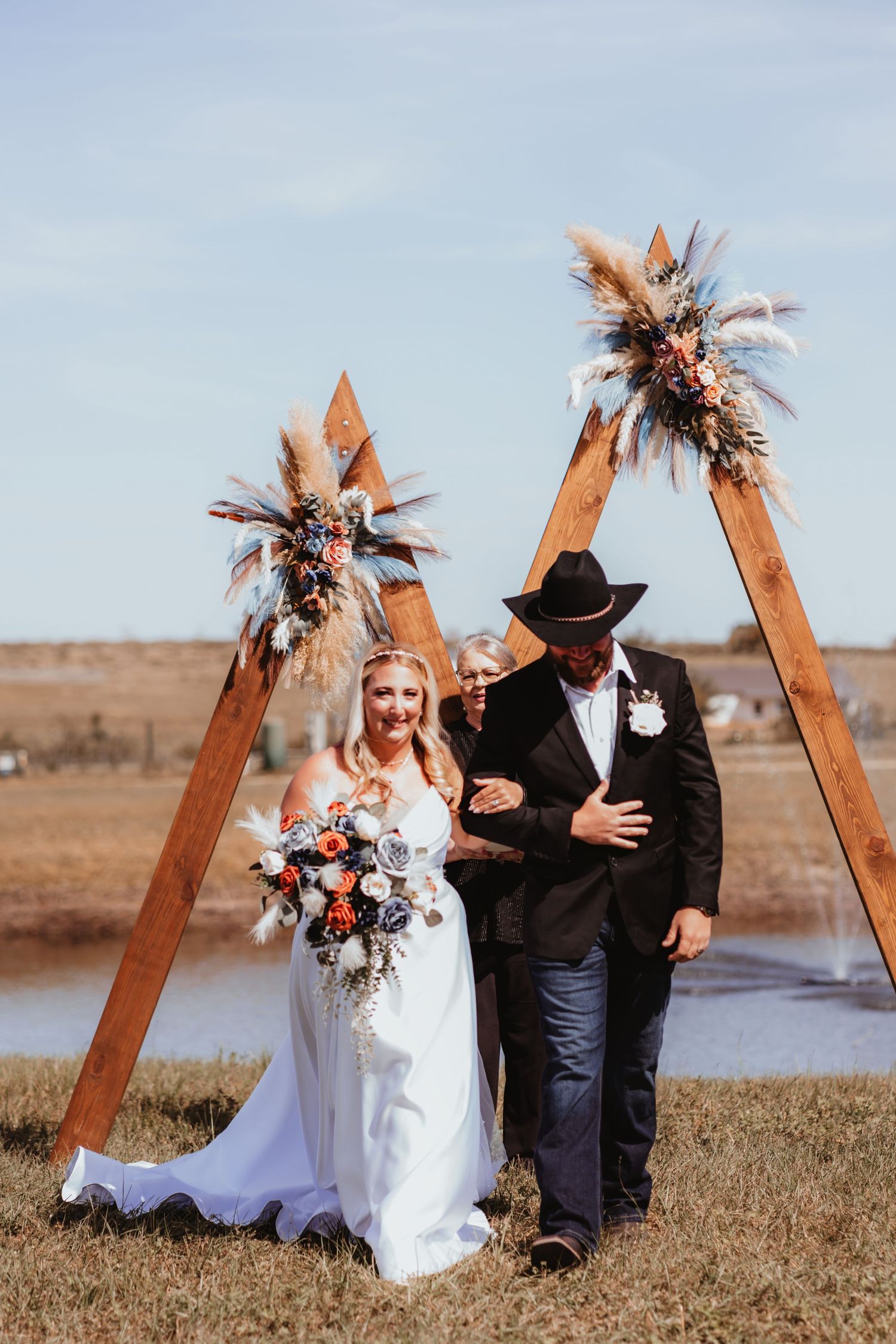 Smiling bride holding bouquet walks with groom in cowboy hat under boho wooden triangle arch at outdoor country wedding.