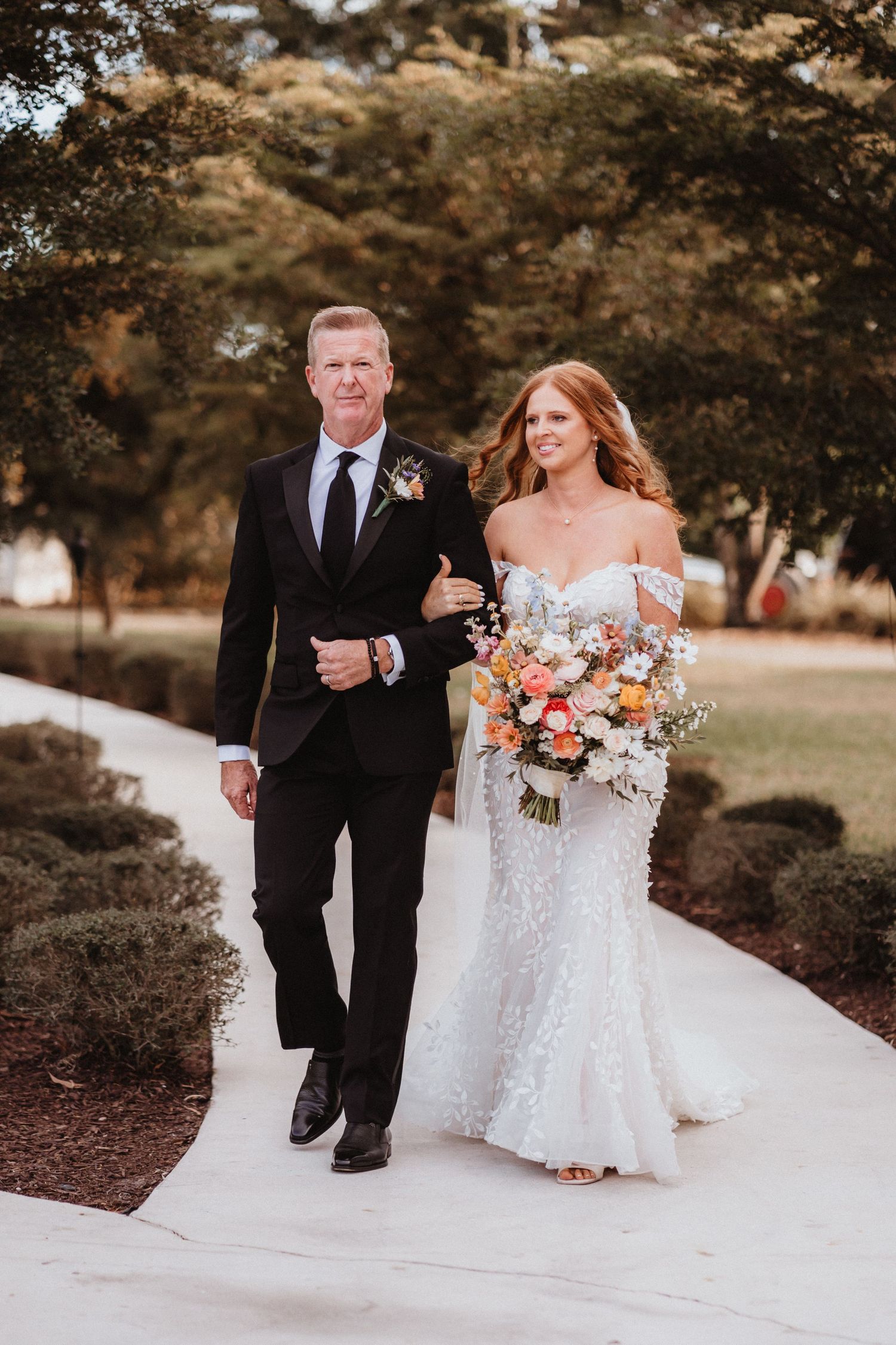 Bride in lace gown holding colorful bouquet walks down aisle with escort in black tuxedo outdoors.