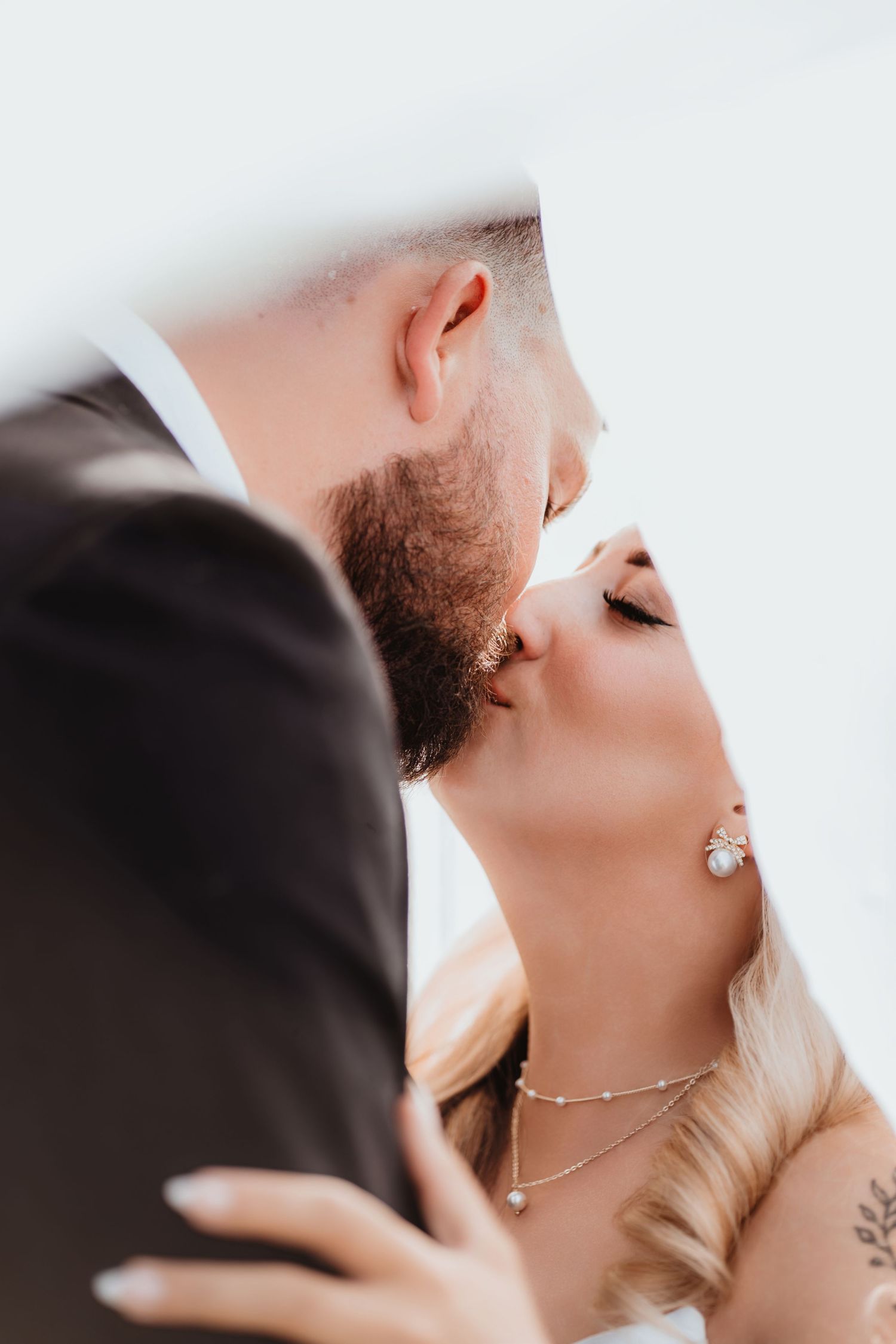 Romantic close-up of bride and groom kissing under a flowing veil, highlighting bride's elegant jewelry and tattoo.