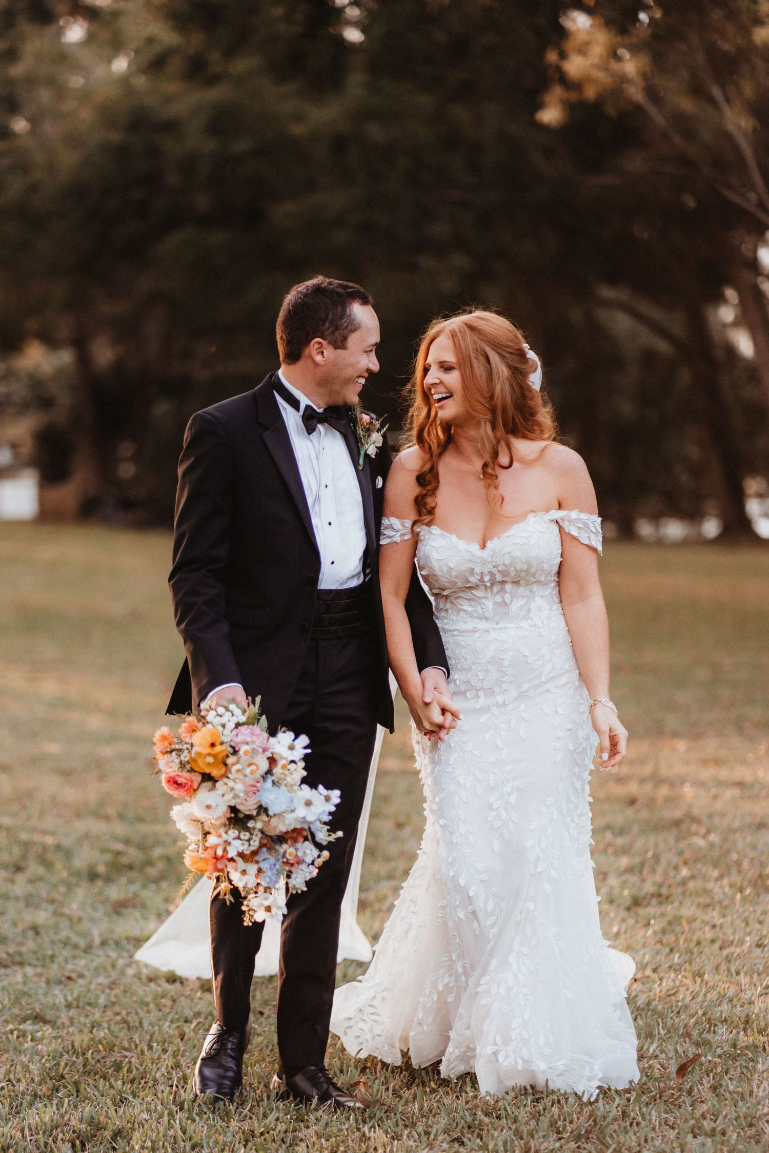 Smiling couple walks through park at sunset, groom holding colorful bouquet, bride in fitted lace off-shoulder gown.