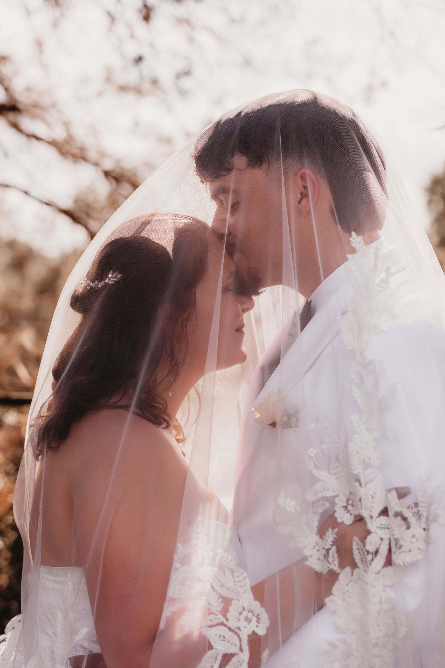 Bride and groom standing close together under a lace-trimmed veil in soft golden sunlight.