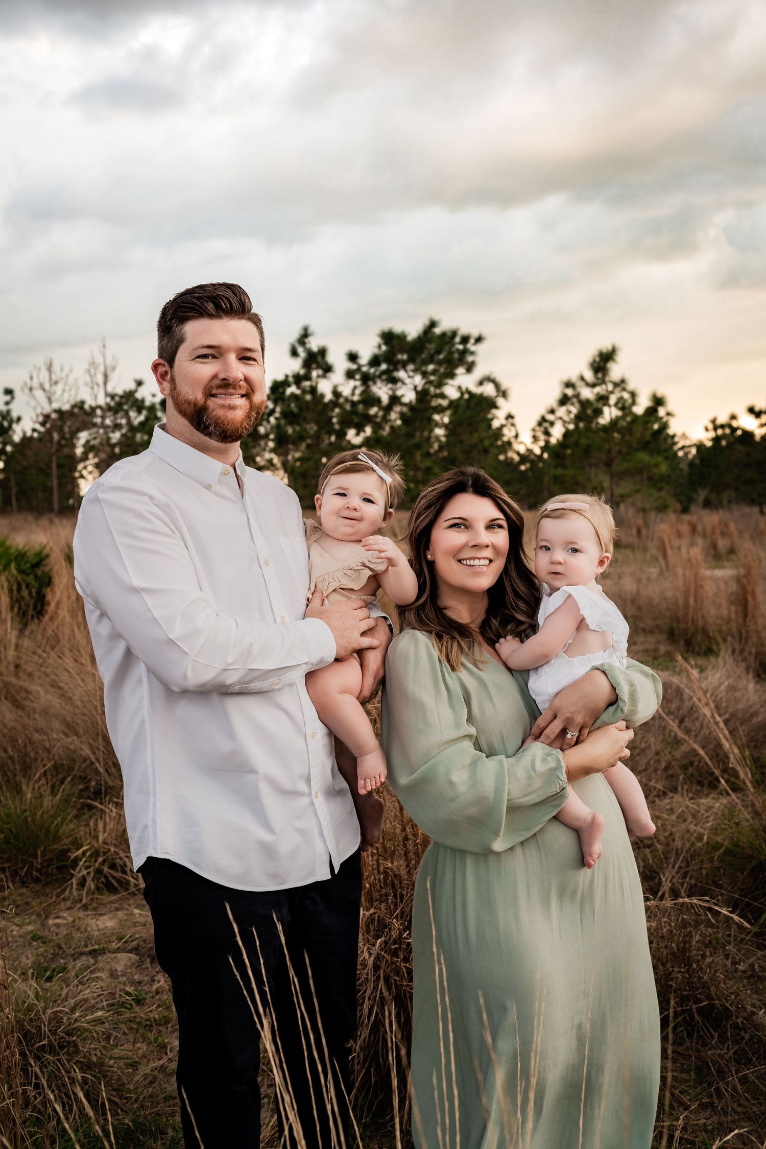 Smiling family of four with two babies posing in a golden field under a dramatic cloudy sky.