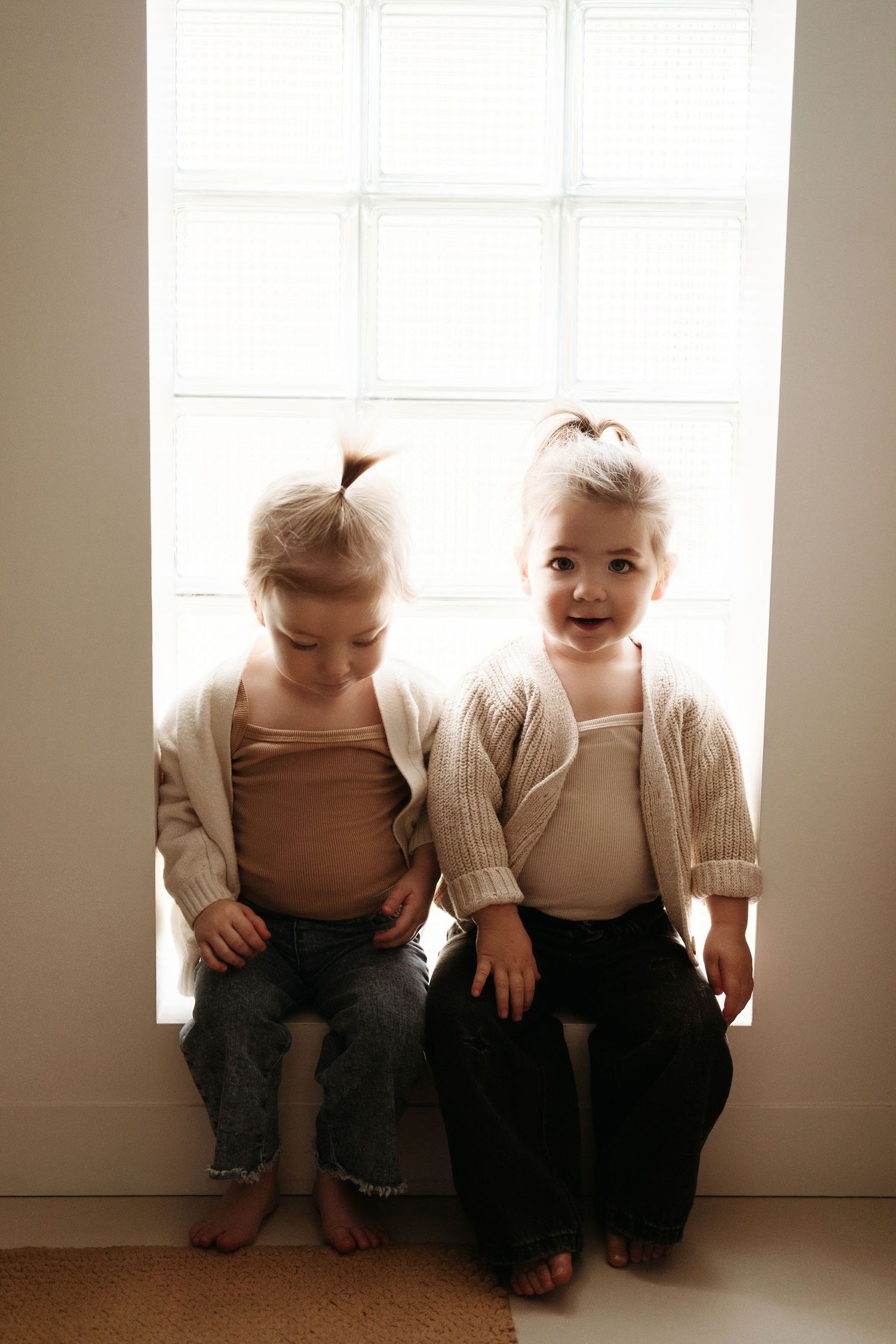 Two young children in matching cream cardigans sit on a bright window sill, one smiling at camera.