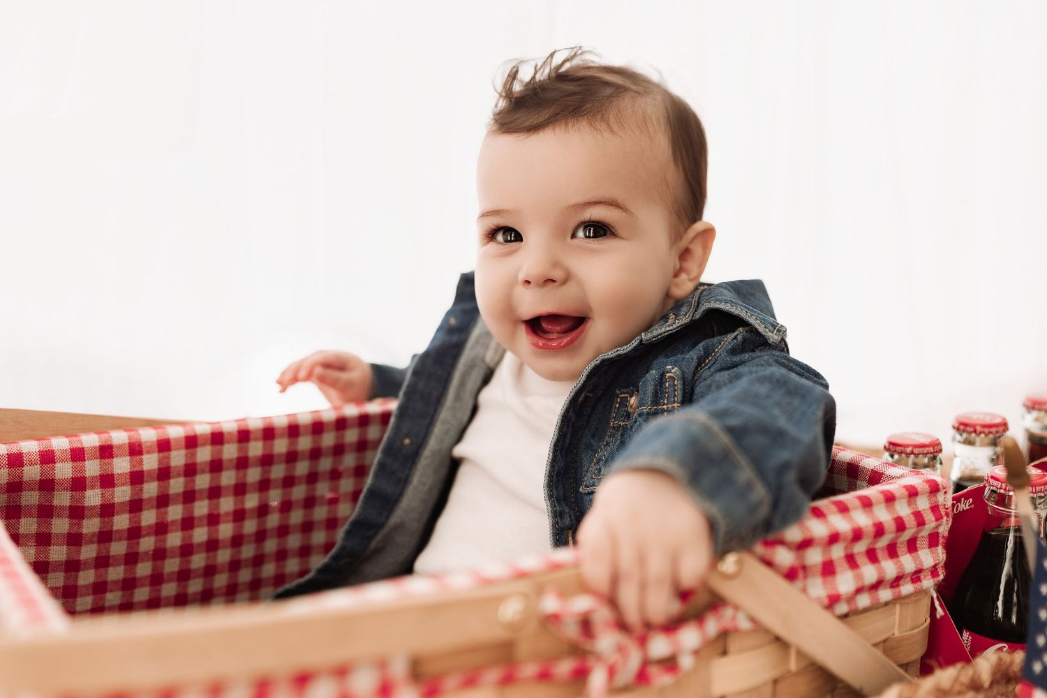 Happy baby in denim jacket laughing joyfully while sitting in a red gingham-lined picnic basket.