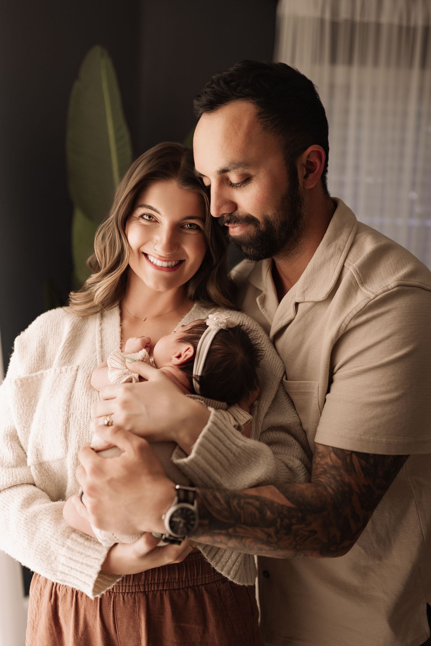 Smiling couple holding newborn baby girl with headband, father with tattoos embracing family.