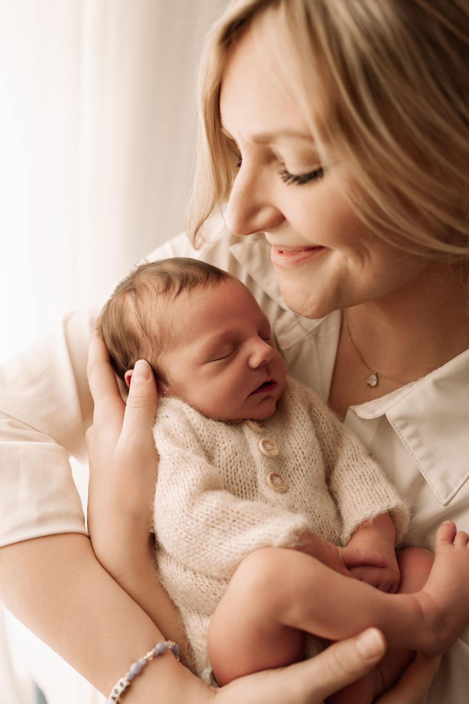 Smiling mother with closed eyes tenderly holding her sleeping newborn in a cream knit outfit.