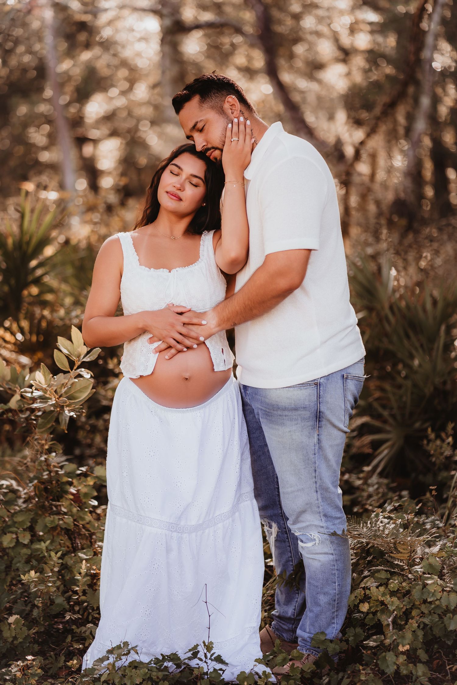 Intimate maternity portrait of couple standing together in woodland, gently holding pregnant belly.
